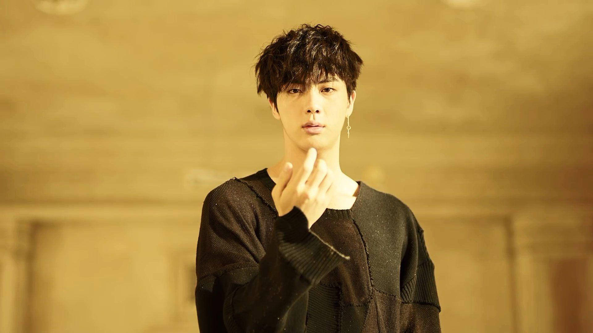 BTS 桌布 壁紙 — 一位黑髮、身穿黑色毛衣的年輕男子，對著溫暖的金色紋理牆壁做出手指愛心手勢。