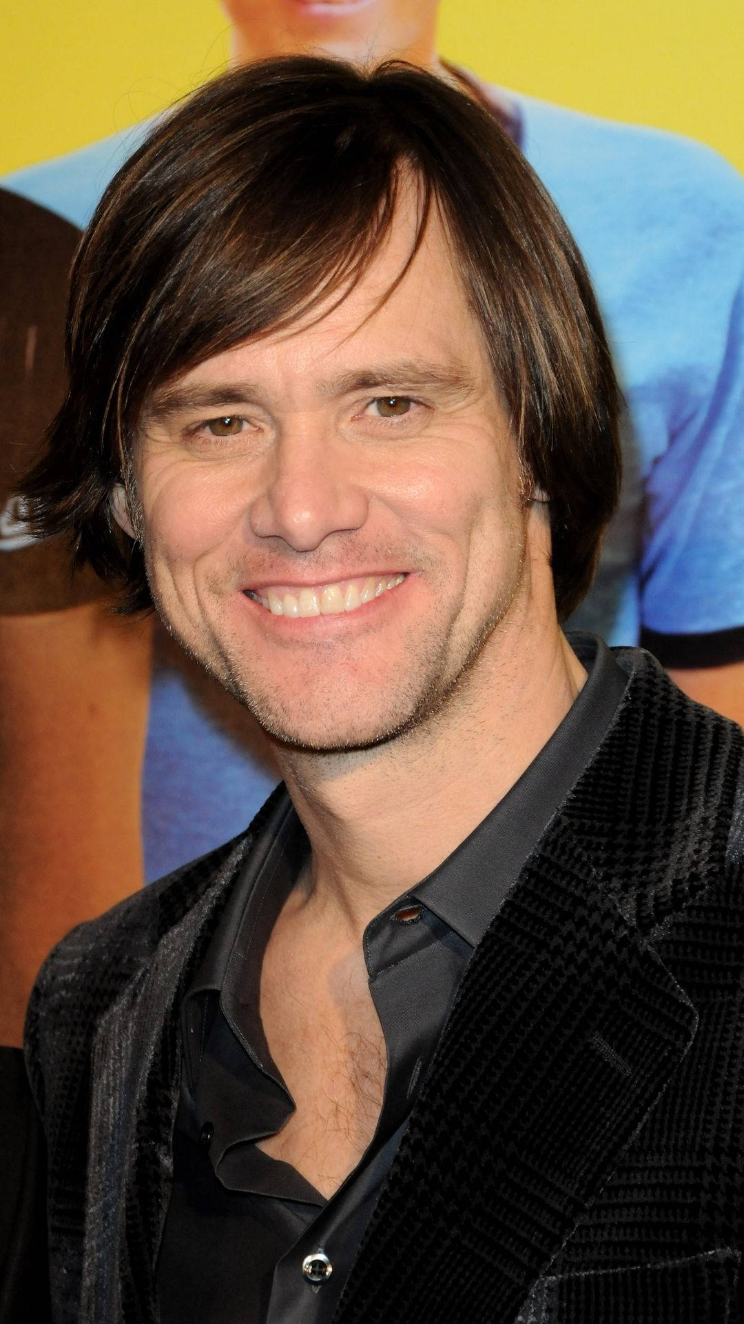 Jim Carrey 桌布 壁紙 — 一位黑髮、身穿黑色紋理外套的微笑男士，在色彩繽紛的模糊背景前擺姿勢。