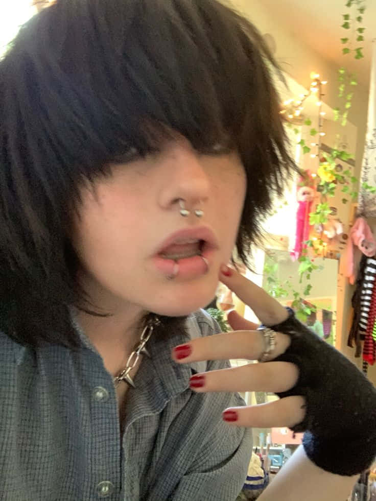 emo 美學 桌布 壁紙 — 一位黑髮且有多處面部穿孔的人，在溫暖照明的房間裡，掛著植物，擺出姿勢，展示紅色指甲和無指手套。
