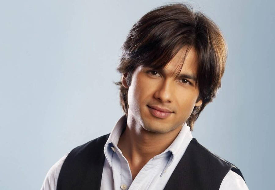 Shahid Kapoor 桌布 壁紙 — 一位黑髮俊男帶著溫柔的微笑，在柔和的藍色背景前，穿著淺藍色襯衫外搭黑色背心。