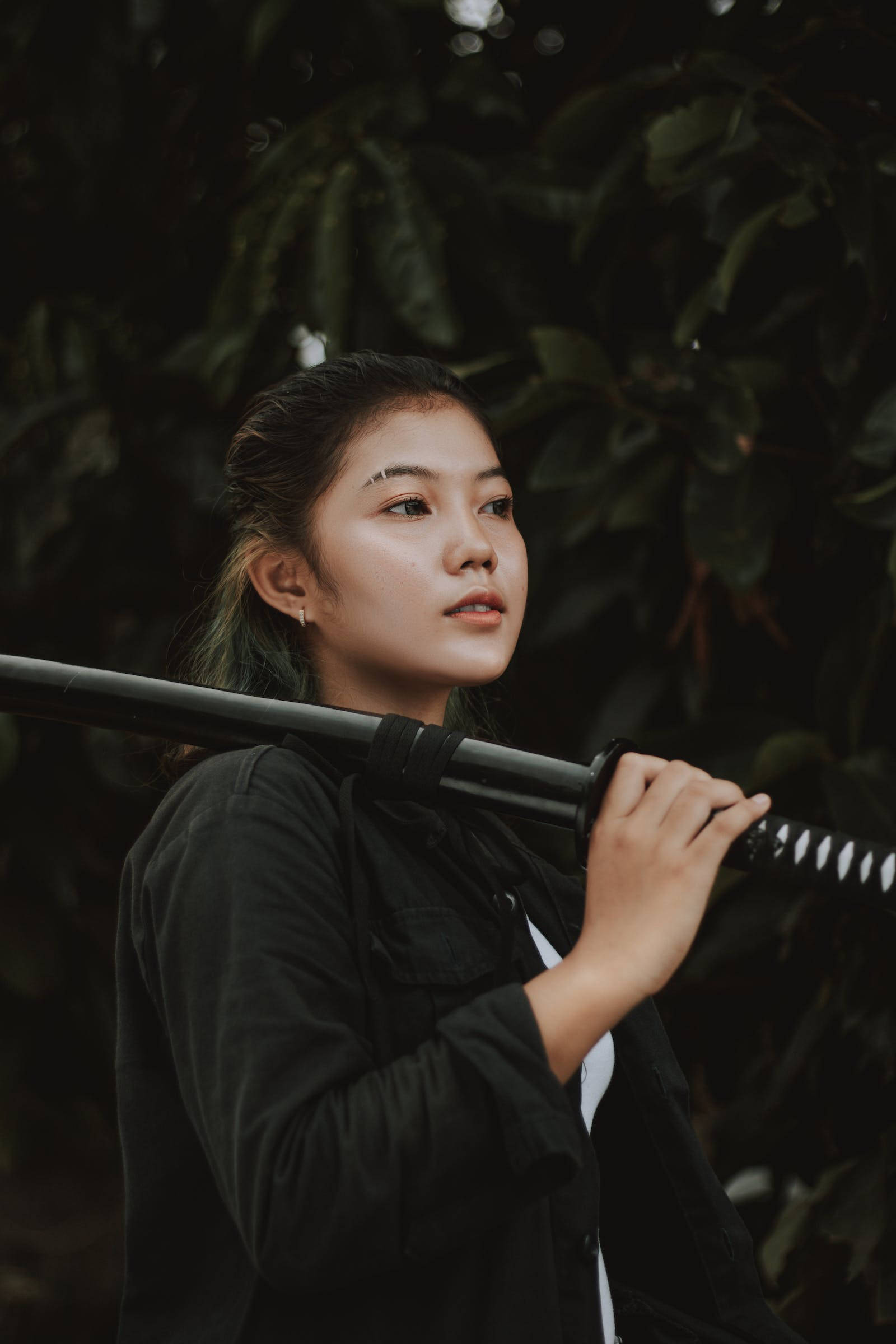 桌布 壁紙 — 一位黑髮年輕女子在茂密綠葉背景下將武士刀扛在肩上。