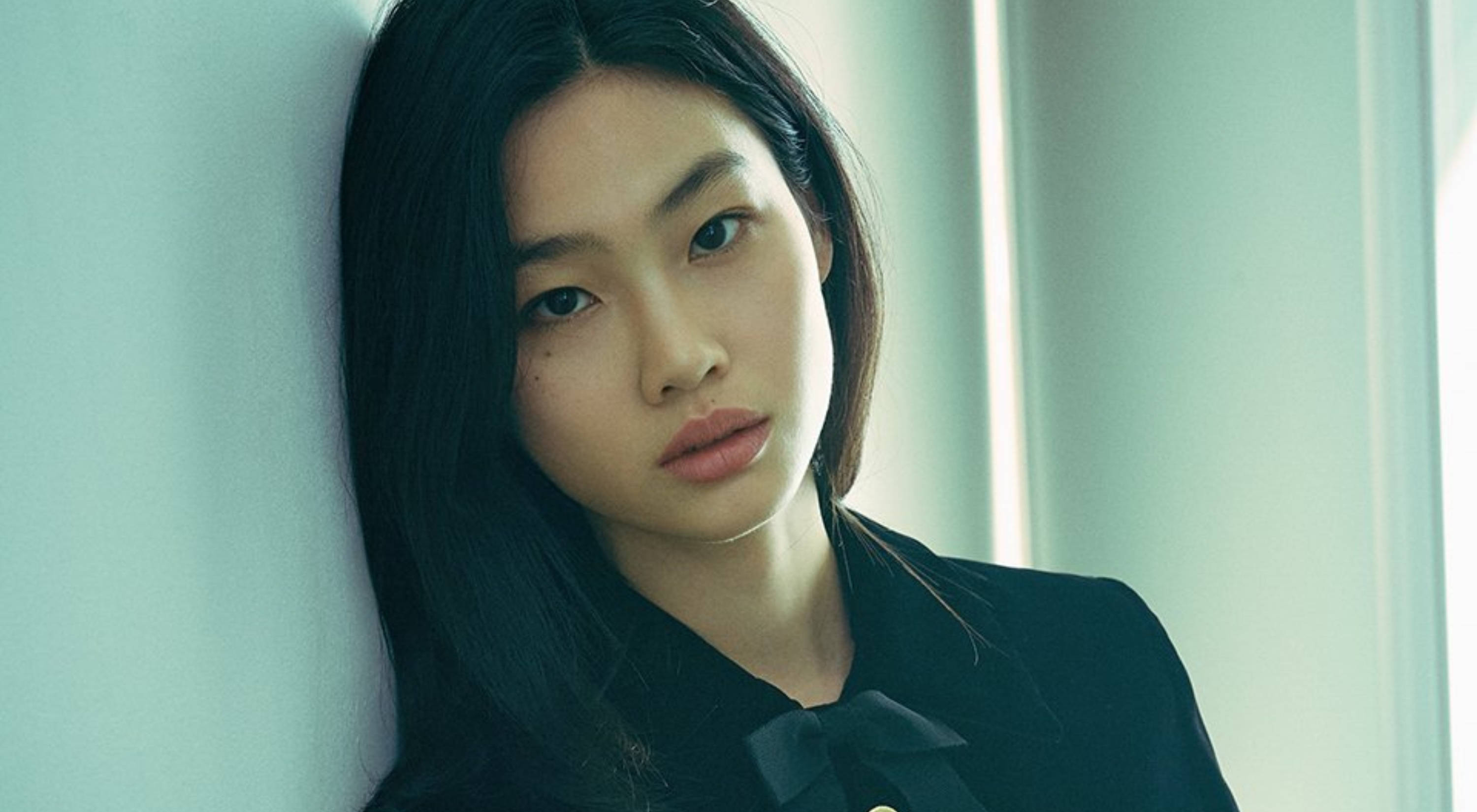 Hoyeon 桌布 壁紙 — 一位黑髮年輕女子靠著淡藍色牆壁，在柔和燈光下專注凝視，突顯她的五官。