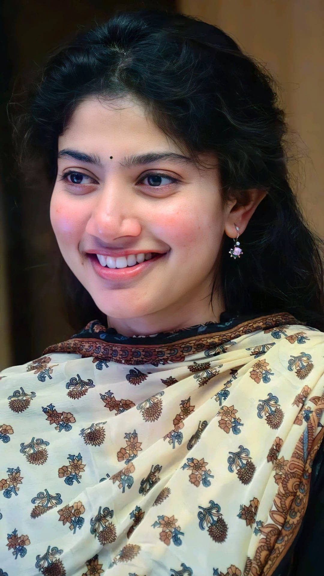 Sai Pallavi 桌布 壁紙 — 一位黑髮年輕女性穿著帶有棕色和藍色花卉圖案的奶油色紗麗，溫暖地微笑。