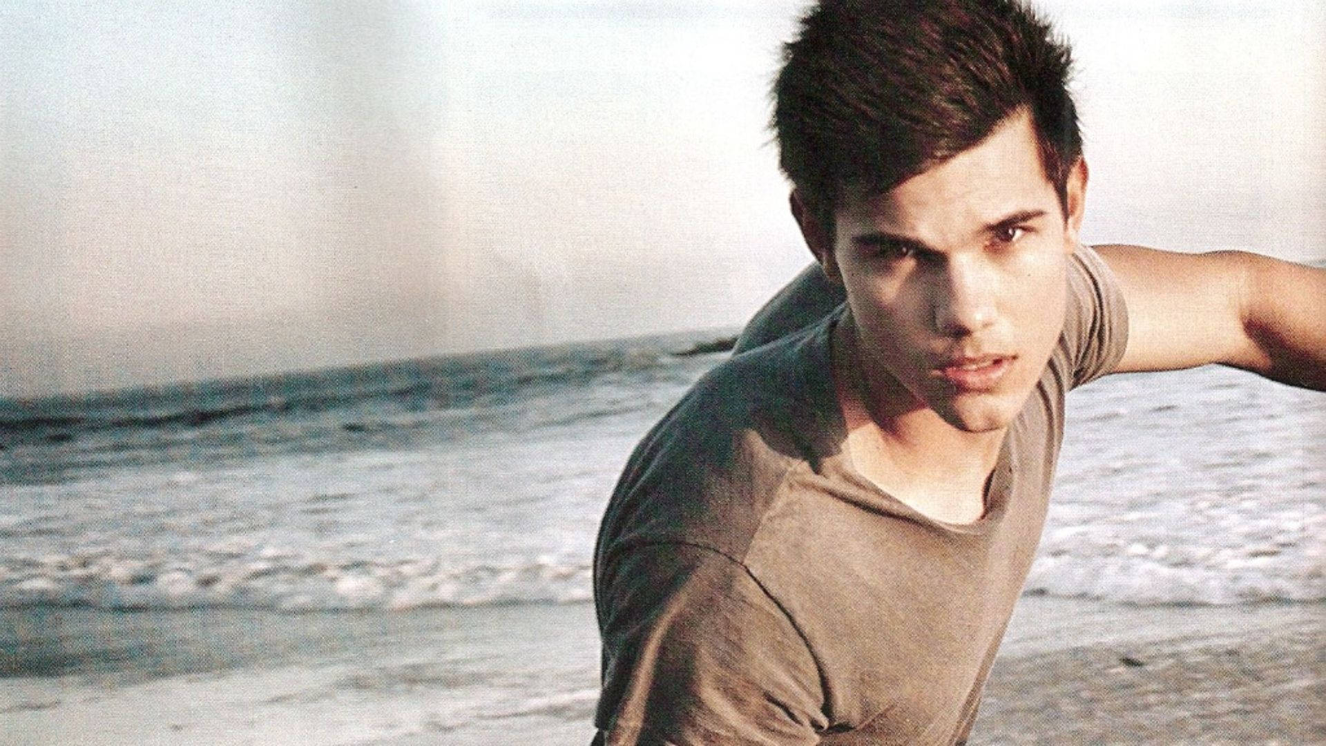 Taylor Lautner 桌布 壁紙 — 一位黑髮年輕男子在沙灘上向前傾，身後有海浪在柔和日光下拍打。