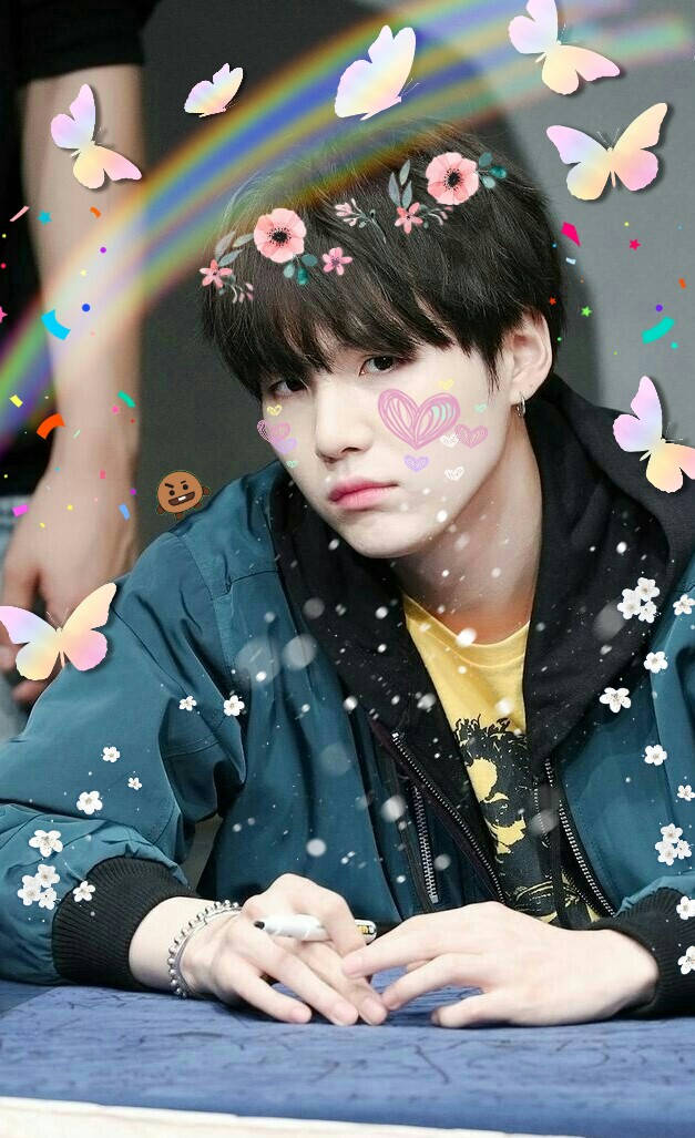 BTS 鎖定螢幕 桌布 壁紙 — 一位黑髮年輕男子坐在桌旁，色彩繽紛的蝴蝶、花朵和彩虹以奇幻的數位疊加效果漂浮在他的頭部周圍。