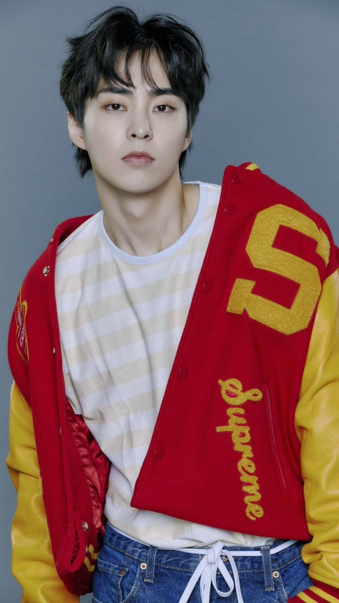 EXO 桌布 壁紙 — 一位黑髮年輕男子身穿紅黃色印有 'Supreme' 品牌的棒球外套，內搭條紋襯衫和牛仔褲，背景為灰色。