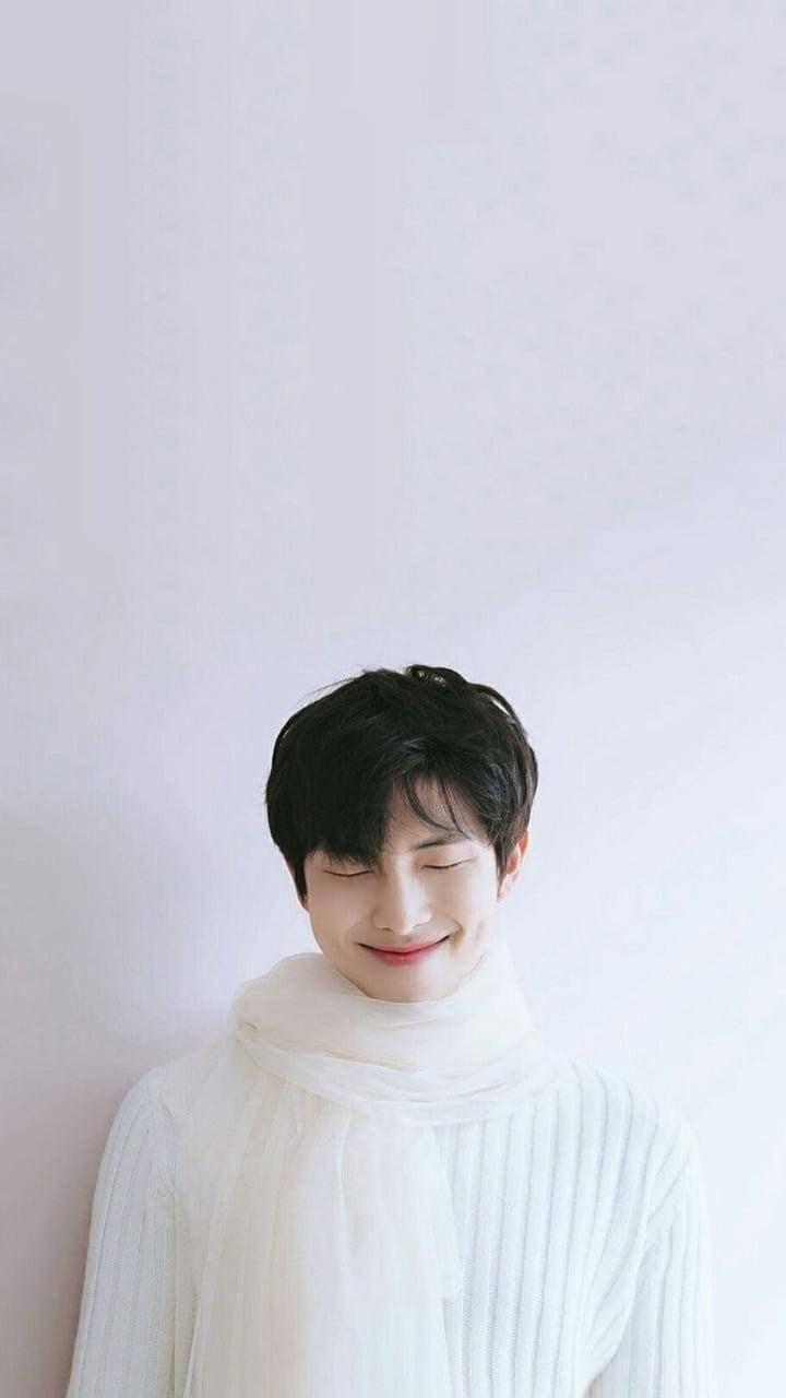 Namjoon 桌布 壁紙 — 一位黑髮年輕男子閉著眼睛溫柔微笑，身穿白色羅紋毛衣和圍巾，背景為純色淺色。