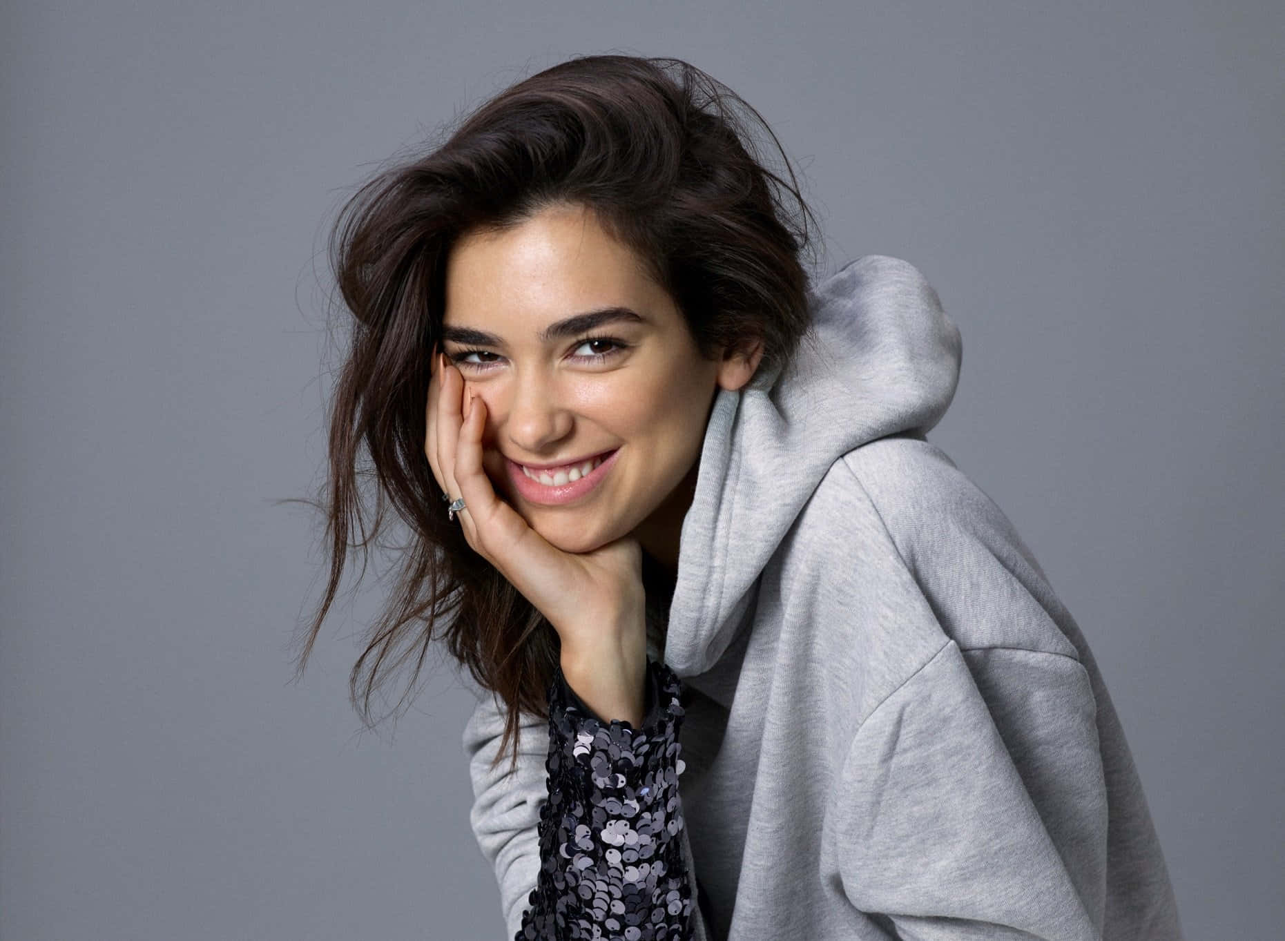 Dua Lipa 桌布 壁紙 — 一位黑髮微笑的女性，身穿灰色連帽衫和亮片上衣，在純灰色背景前將臉頰靠在手上。
