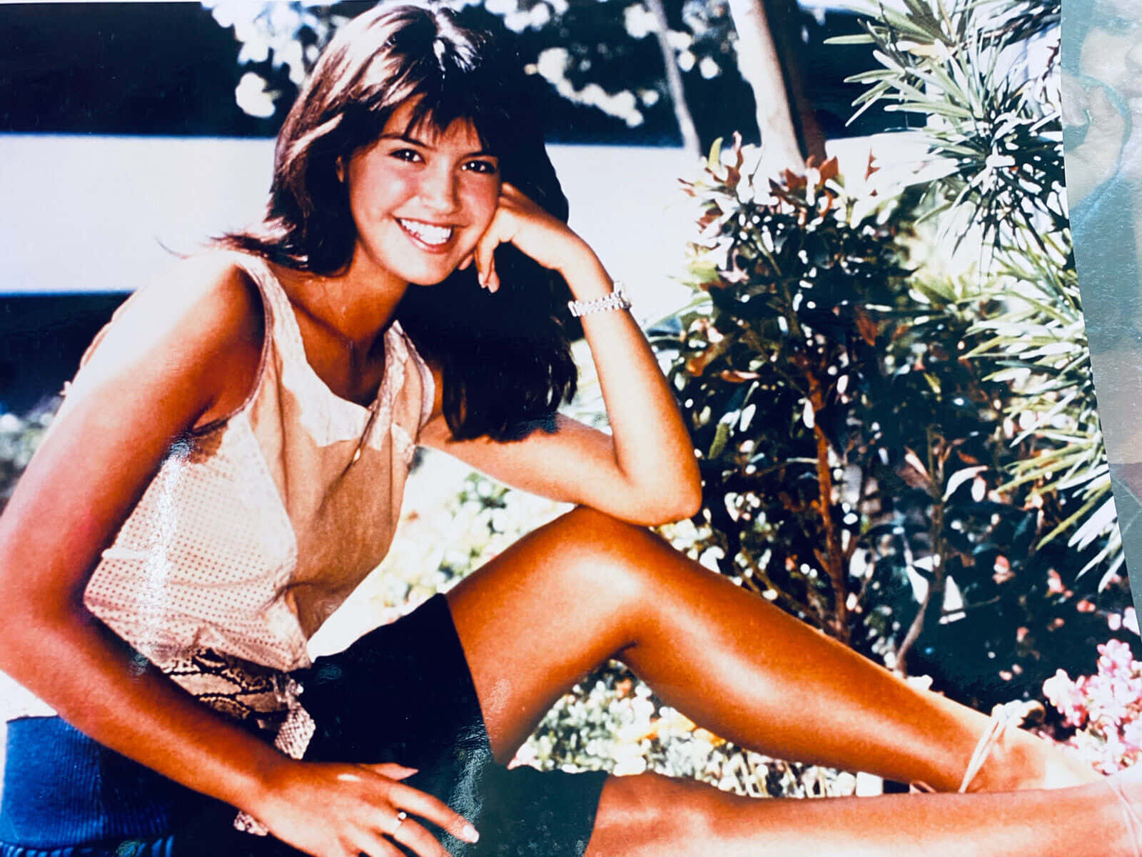 Phoebe Cates 桌布 壁紙 — 一位黑髮微笑的女性坐在戶外明亮的陽光下，身穿淺色上衣和深色短褲，被綠植環繞。