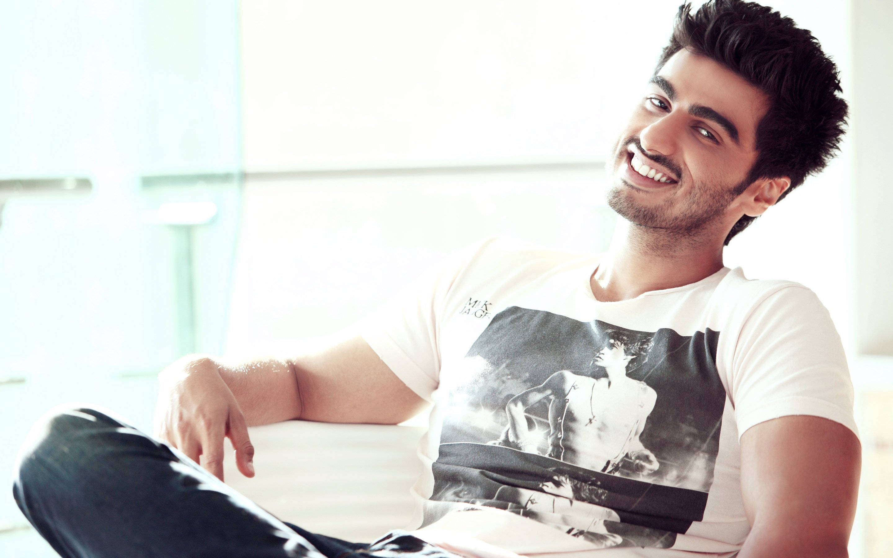 Arjun Kapoor 桌布 壁紙 — 一位黑髮微笑的男子在明亮的現代房間裡，悠閒地躺在白色沙發上。
