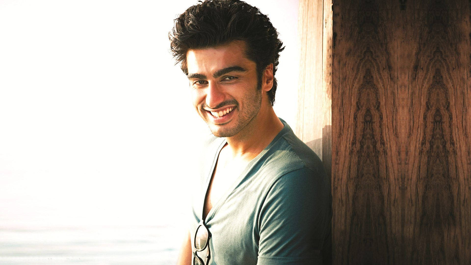 Arjun Kapoor 桌布 壁紙 — 一位黑髮微笑的男子靠在沐浴在溫暖陽光下的木牆上。