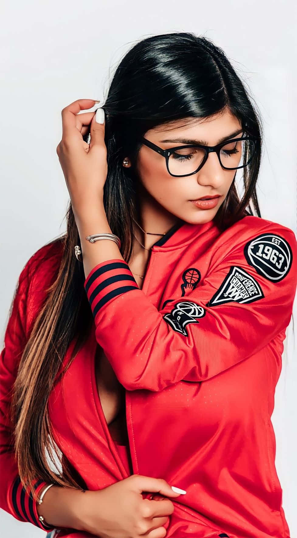 Mia Khalifa 桌布 壁紙 — 一位黑髮戴眼鏡的年輕女性，在純白色背景前穿著一件帶有籃球補丁的鮮紅色轟炸機夾克。