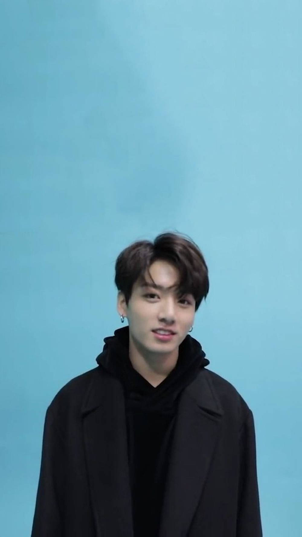 BTS 桌布 壁紙 — 一位黑髮戴耳環的年輕男子，穿著黑色外套，在純藍色背景前溫柔微笑。