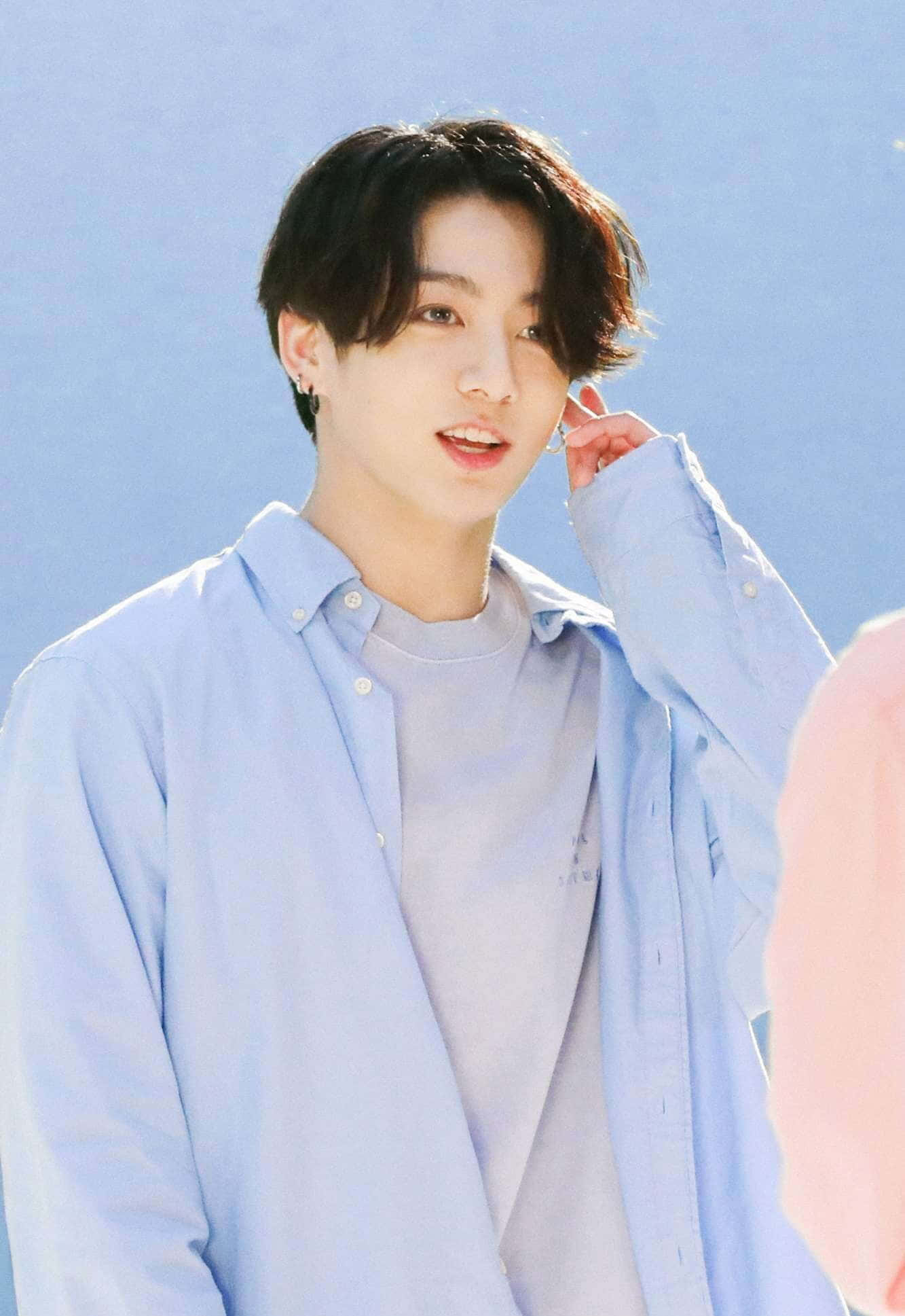 Jungkook 桌布 壁紙 — 一位黑髮戴耳環的年輕男子在清澈的藍天下輕輕微笑，一邊整理他的淺藍色鈕扣襯衫。