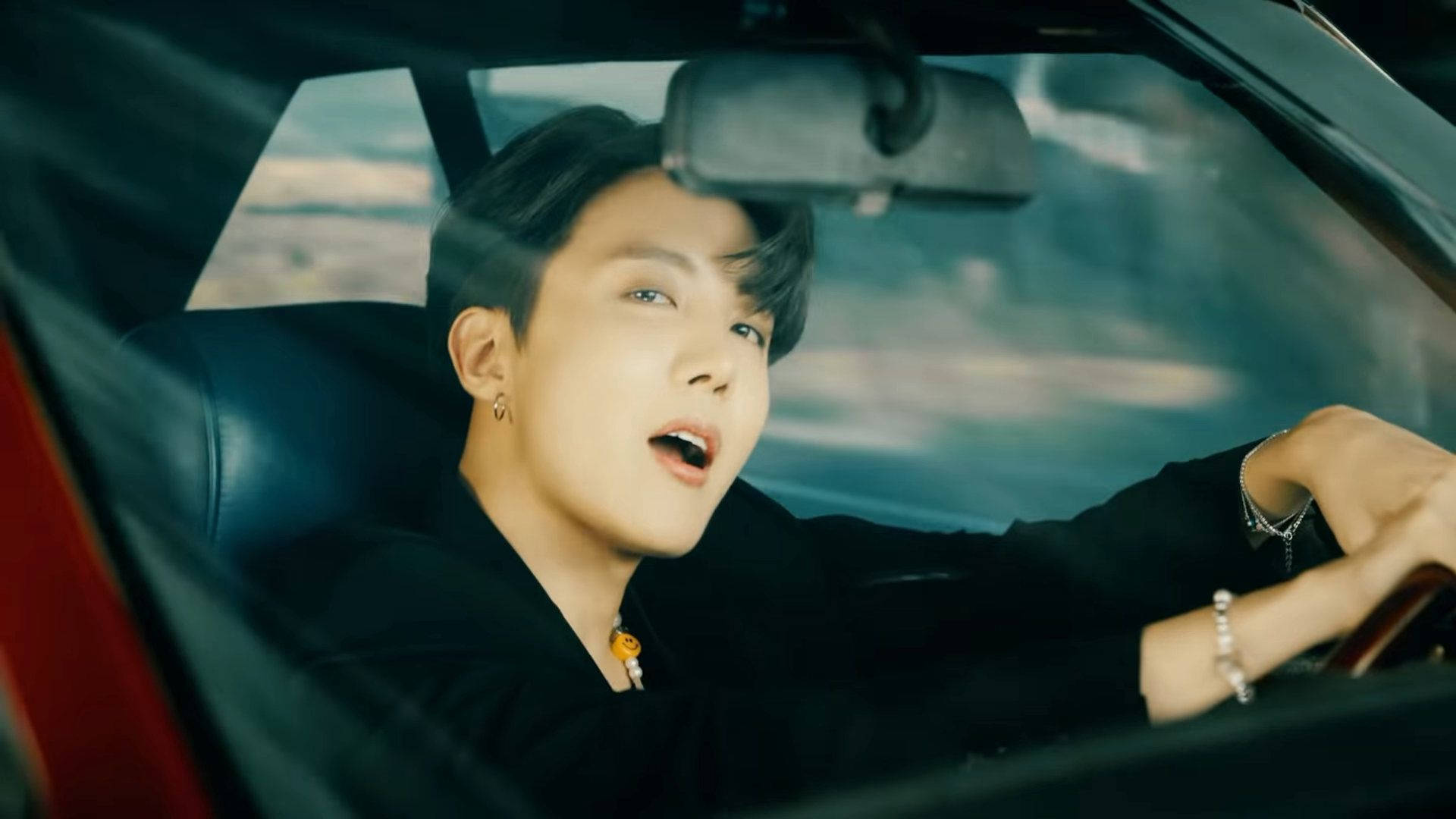 j-hope 桌布 壁紙 — 一位黑髮戴耳環的年輕男子張開嘴開車，身穿黑色衣服和銀色手鐲。