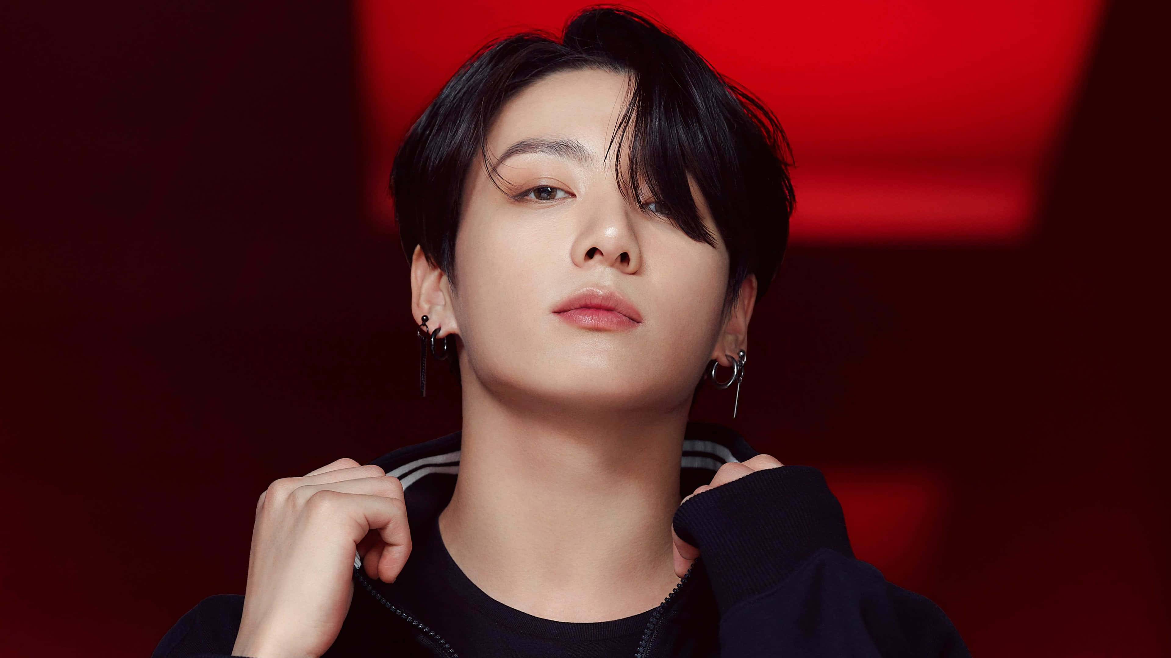 BTS 桌布 壁紙 — 一位黑髮戴著圓環耳環的年輕男子，在深紅色模糊背景前向上凝視，同時調整他的黑色連帽衫。