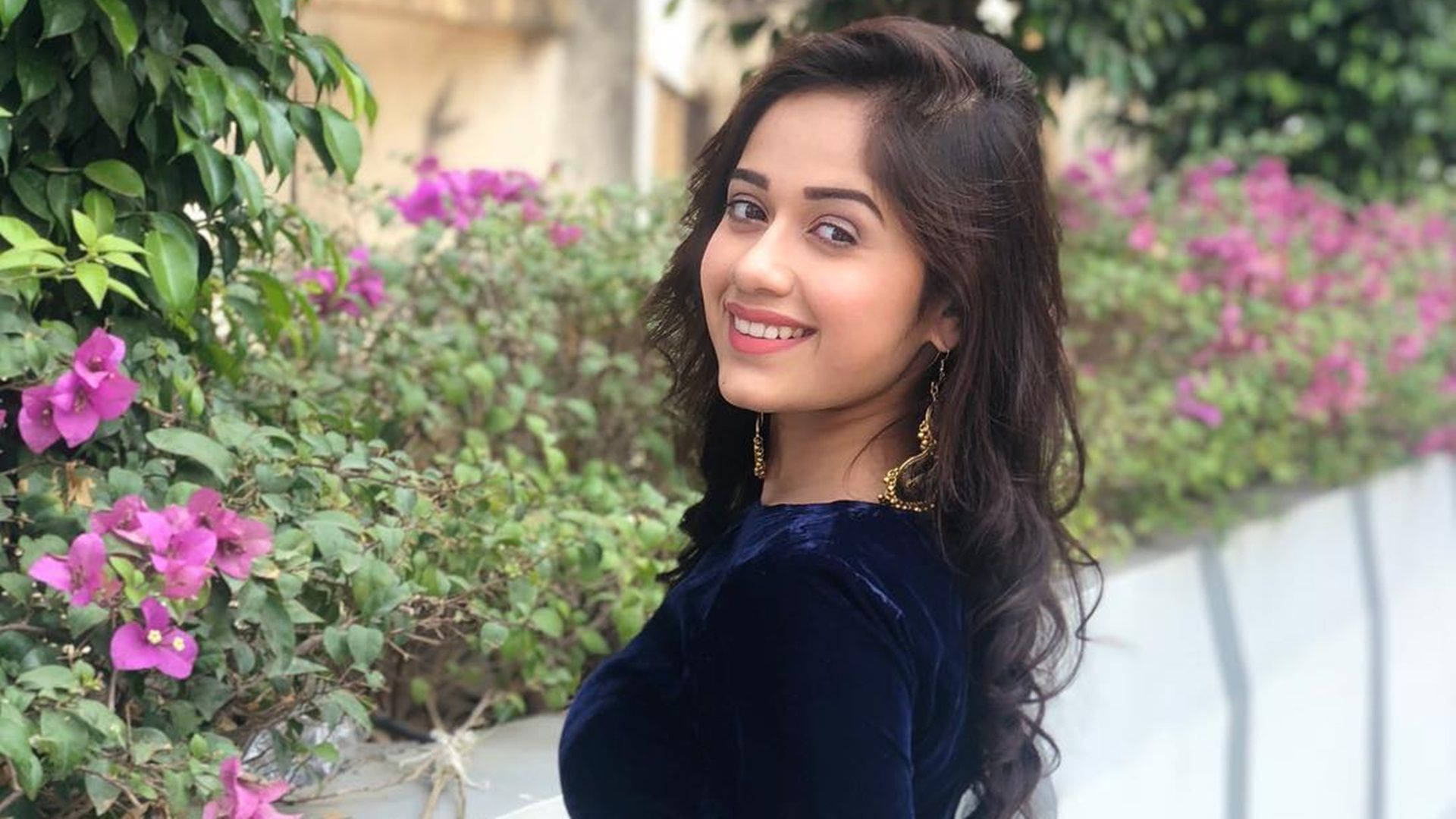 Indian 桌布 壁紙 — 一位黑髮戴金耳環的微笑女士，身穿海軍藍天鵝絨上衣，背景是茂密的綠植和鮮豔的粉紅色九重葛。