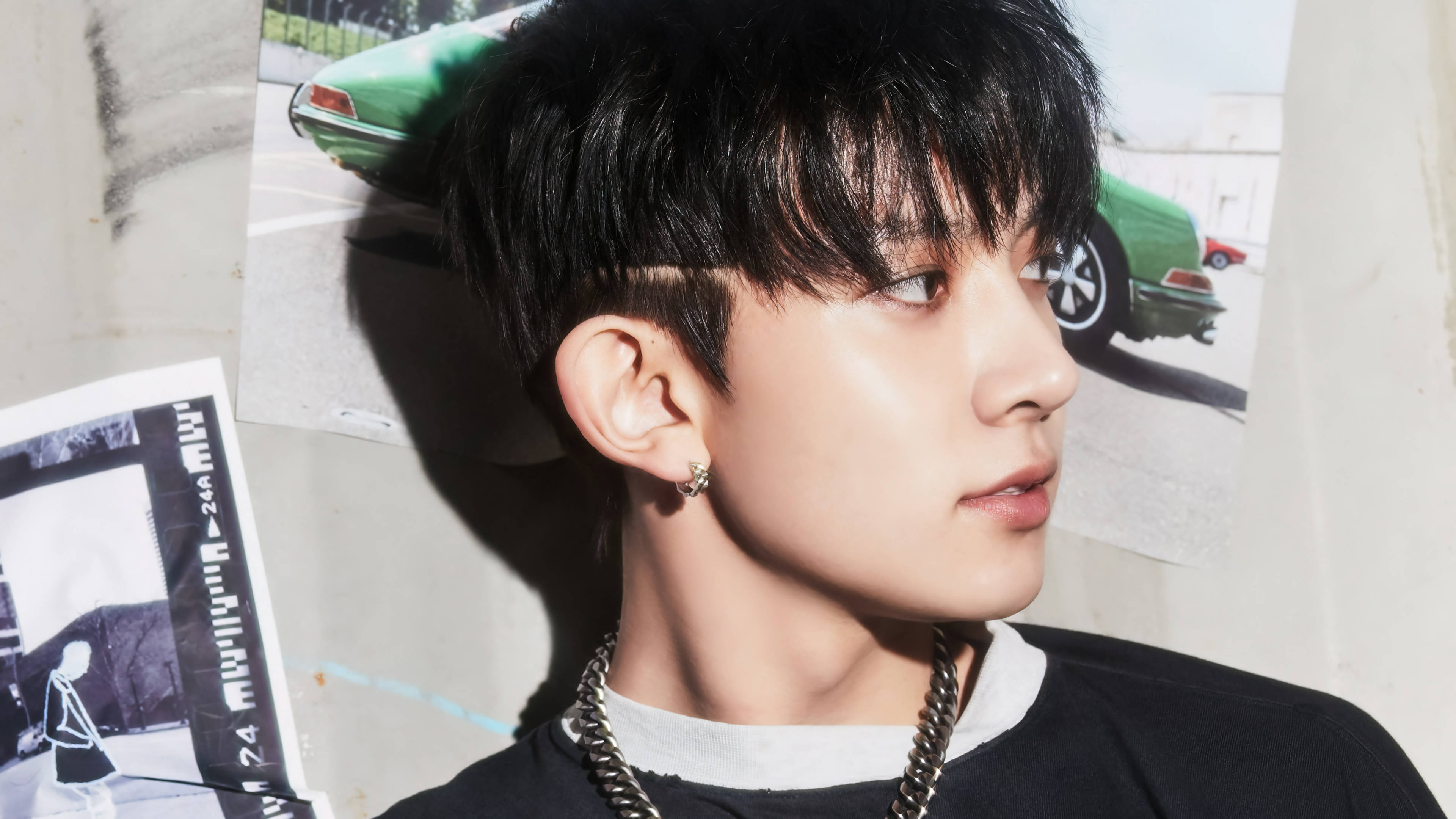 Kpop Aesthetic 桌布 壁紙 — 一位黑髮戴鏈項鍊的年輕男子在質感牆上的復古汽車海報旁若有所思地凝視。