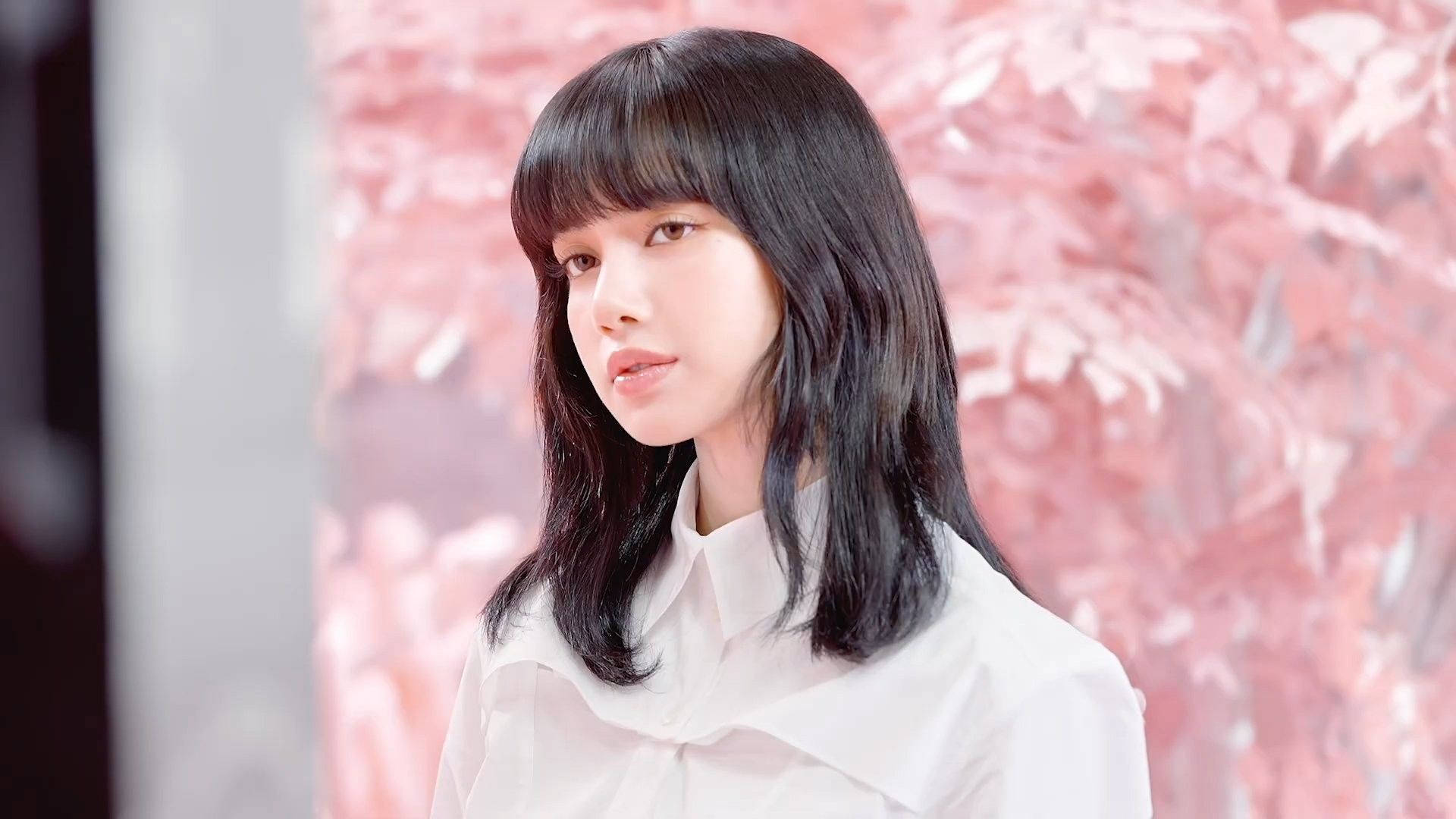 Lalisa 桌布 壁紙 — 一位黑髮有瀏海的年輕女性，身穿白色襯衫，背景是柔和的粉紅色花卉壁紙。