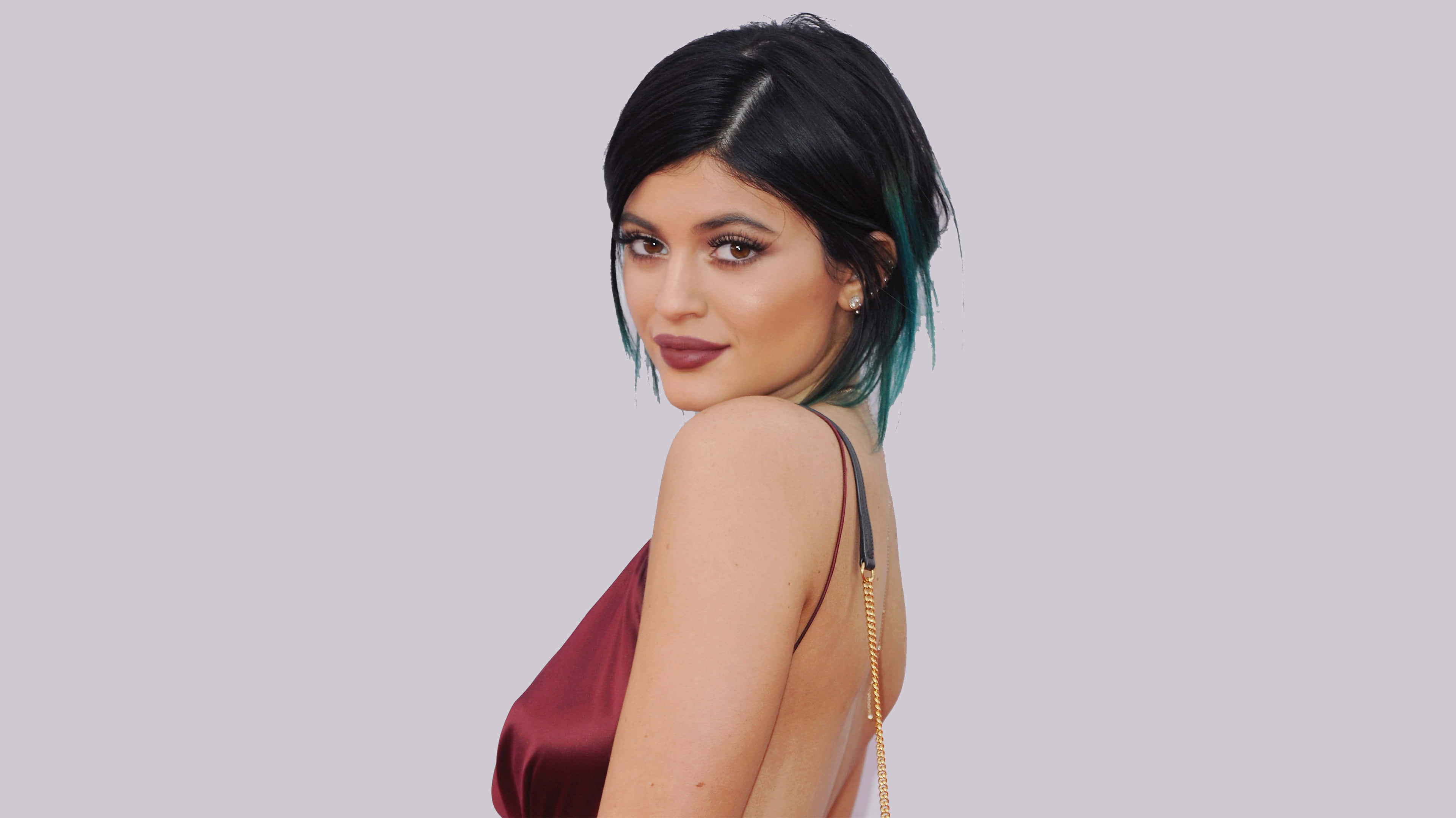 Kylie Jenner 4K 桌布 壁紙 — 一位黑髮末端染成藍綠色的女性，身穿酒紅色緞面上衣，在純灰色背景上轉頭越過肩膀。