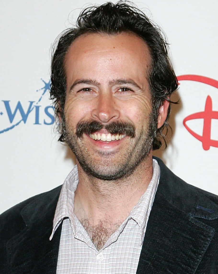 Jason Lee 桌布 壁紙 — 一位黑髮濃鬍鬚的男士微笑著，在品牌背景前穿著深色外套配格紋襯衫。