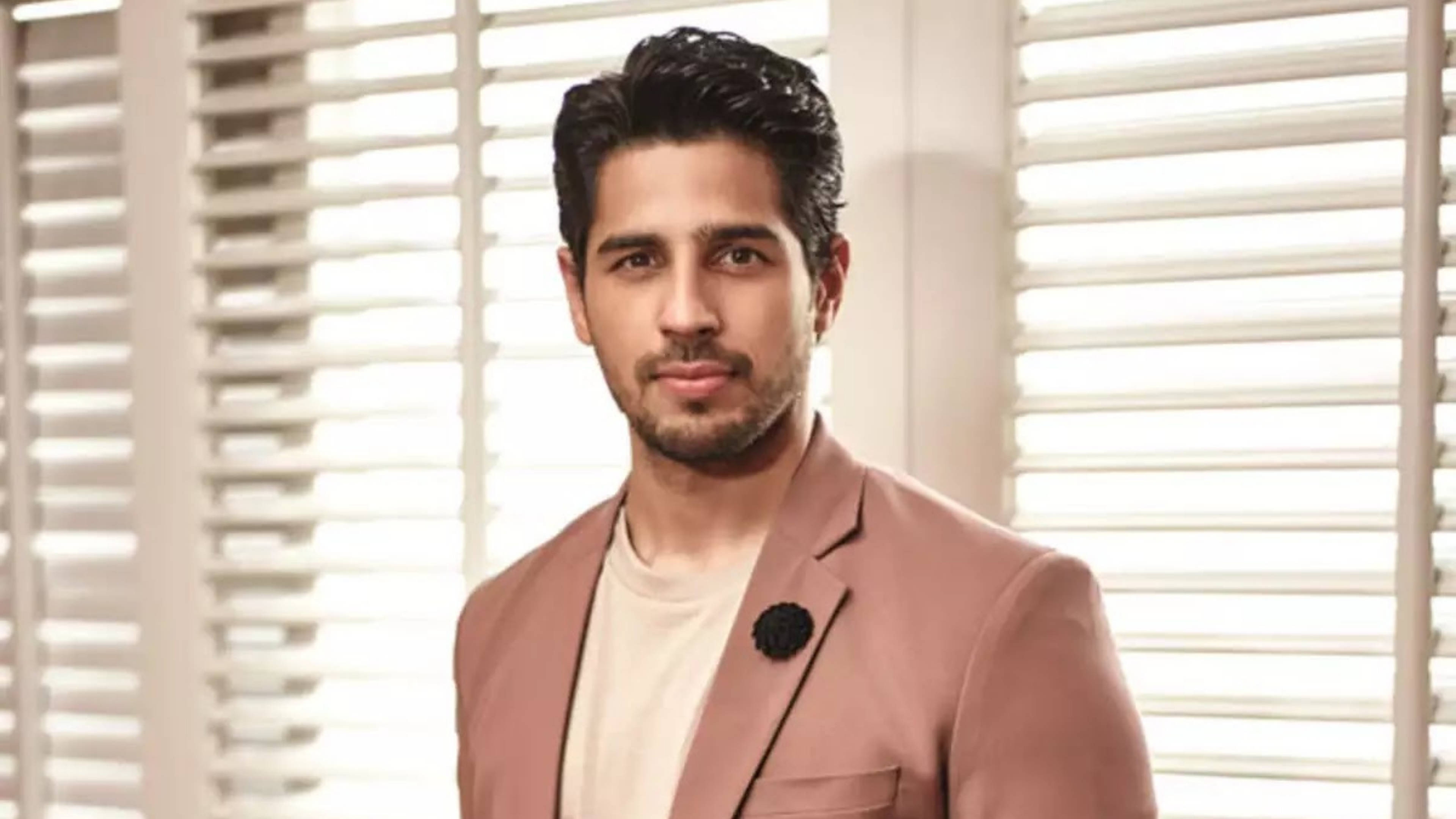 Sidharth Malhotra 桌布 壁紙 — 一位黑髮留鬍的男子，身穿粉紅色西裝外套，內搭奶油色襯衫，別著黑色花朵胸針，背景是白色百葉窗。