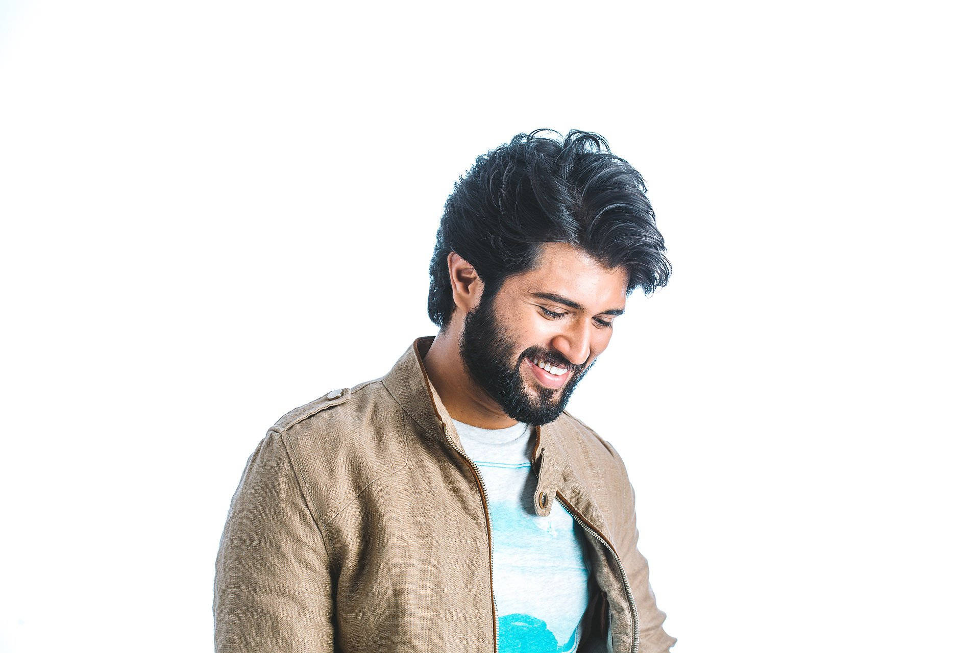 vijay devarakonda 4K 桌布 壁紙 — 一位黑髮留鬍鬚的男士面帶微笑，身穿藍色襯衫外搭棕褐色外套，背景為純白色。