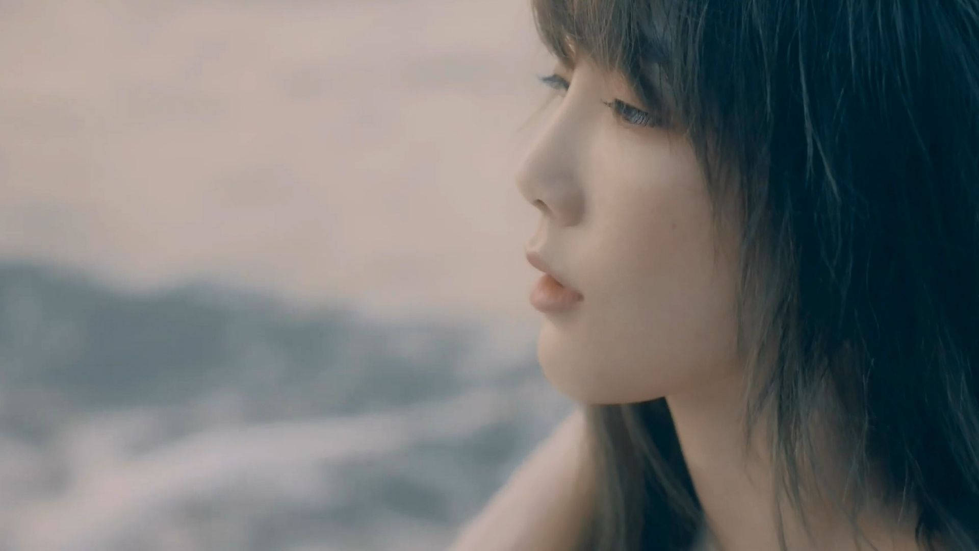 Taeyeon 桌布 壁紙 — 一位黑髮的寧靜女性在柔和散射光線下，若有所思地凝視著模糊的海岸地平線。