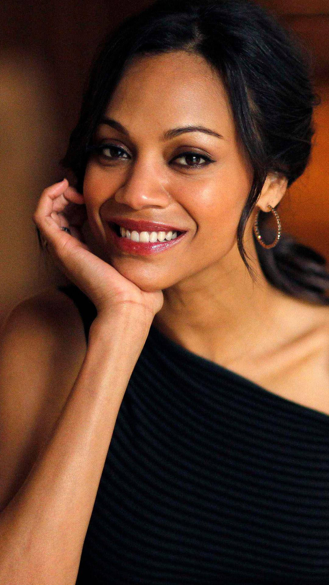 Zoe Saldana 桌布 壁紙 — 一位黑髮的燦爛女性，在柔和的模糊背景中，將下巴擱在手上，溫暖地微笑。