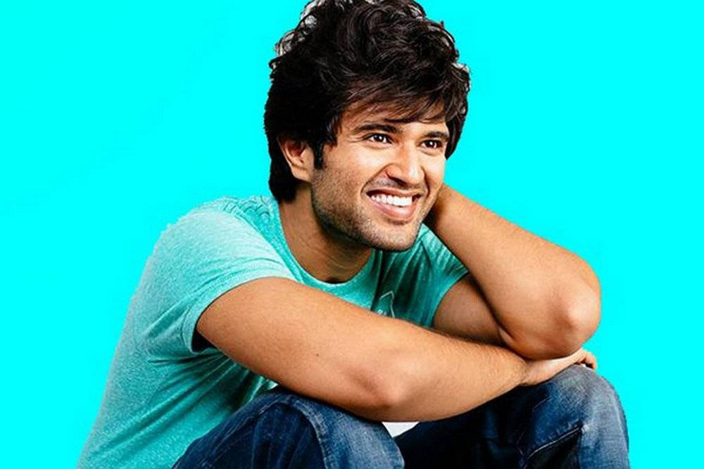 vijay devarakonda 4K 桌布 壁紙 — 一位黑髮的開朗年輕男子，在明亮的青色背景上靠著手臂，露出溫暖的笑容。