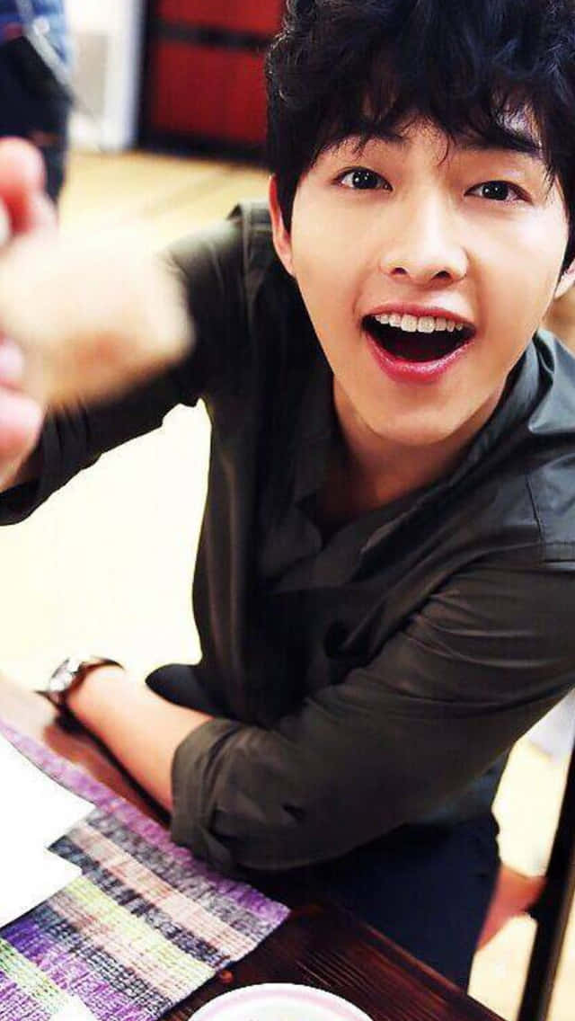 Song Joong Ki 桌布 壁紙 — 一位黑髮的開朗年輕男子在木桌上舉起糕點，露出燦爛的笑容。