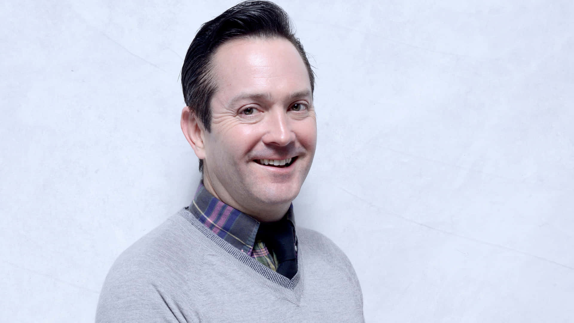 thomas lennon 桌布 壁紙 — 一位黑髮的開朗男士穿著灰色毛衣內搭格紋襯衫，在淺色紋理牆前露出溫暖的笑容。