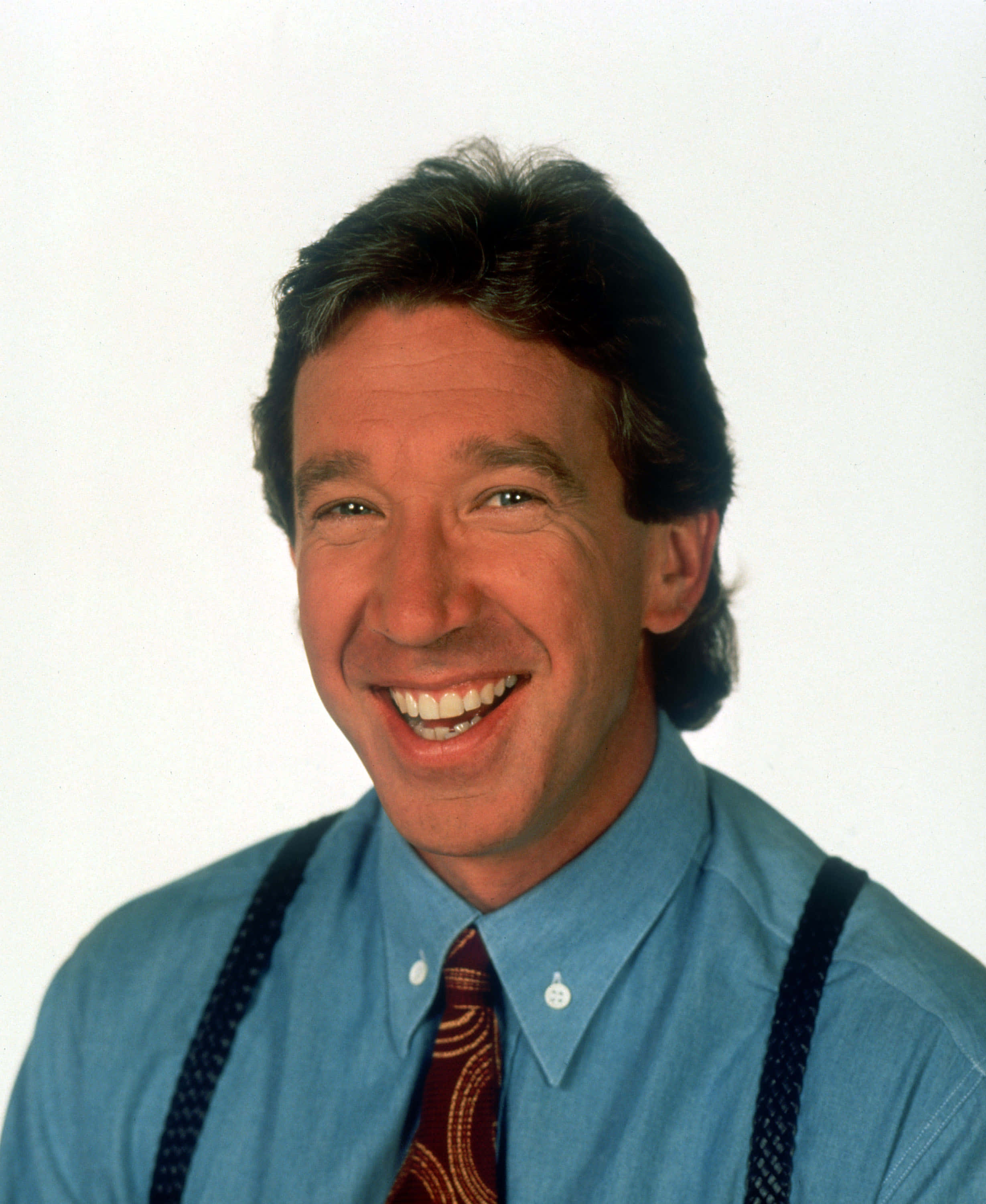 Tim Allen 桌布 壁紙 — 一位黑髮的開朗男子，穿著藍色襯衫、吊帶褲和圖案領帶，在純白背景上燦爛微笑。