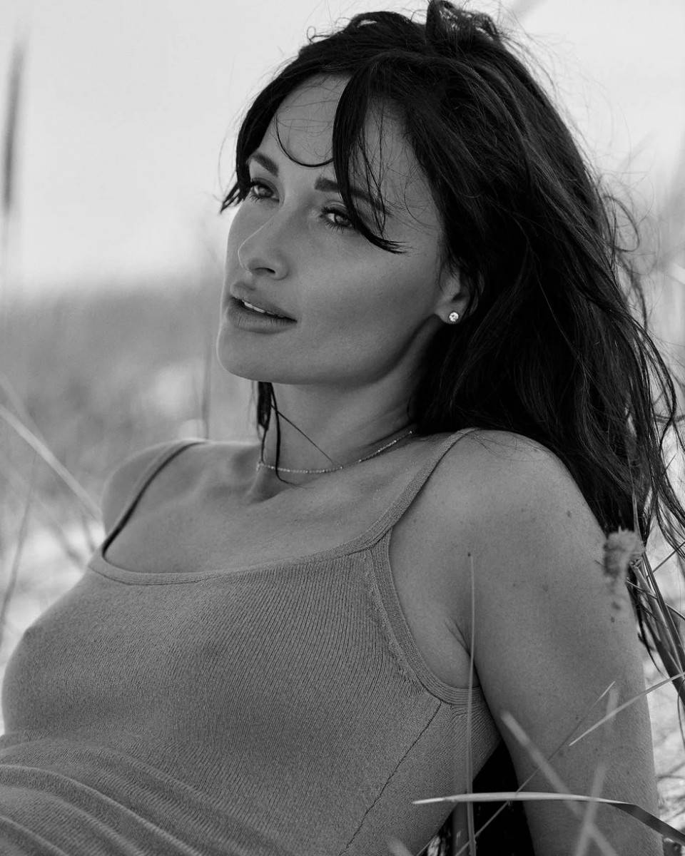 Kacey Musgraves 桌布 壁紙 — 一位黑髮穿背心的女性在高草叢中若有所思地凝視的黑白肖像。