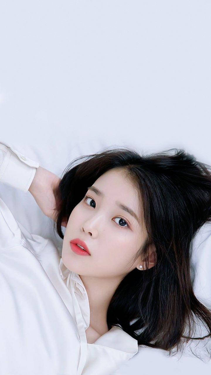 iu phone 桌布 壁紙 — 一位黑髮紅唇的年輕女子躺在白色床上，以柔和的表情抬頭看著相機。