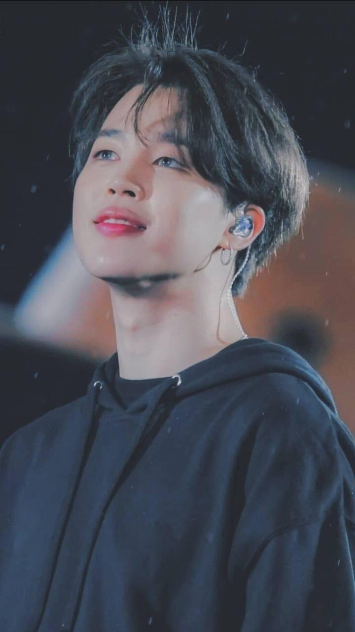 Jimin Aesthetic 桌布 壁紙 — 一位黑髮紅唇的年輕男子在舞台燈光下穿著黑色連帽衫和耳機，向上凝視。