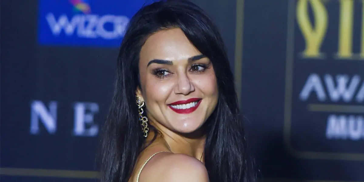 Preity Zinta 桌布 壁紙 — 一位黑髮紅唇的微笑女性在帶有品牌背景的音樂頒獎活動中擺姿勢。
