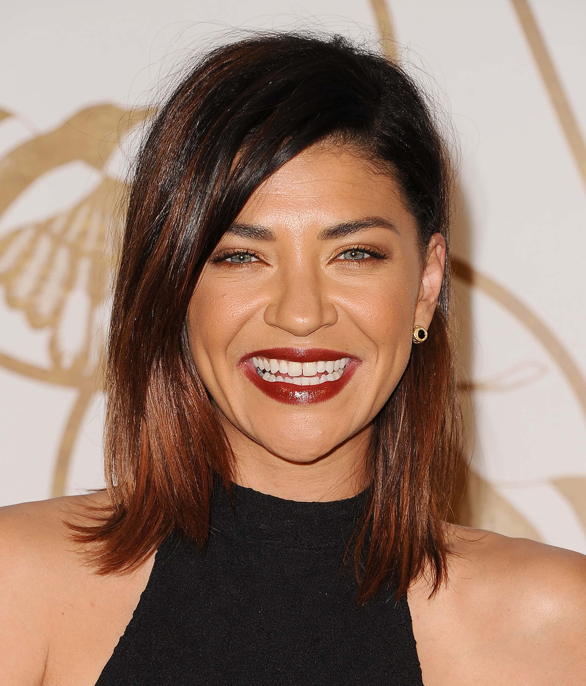 jessica szohr 桌布 壁紙 — 一位黑髮紅唇的耀眼女性，在金色徽章背景前燦爛微笑。