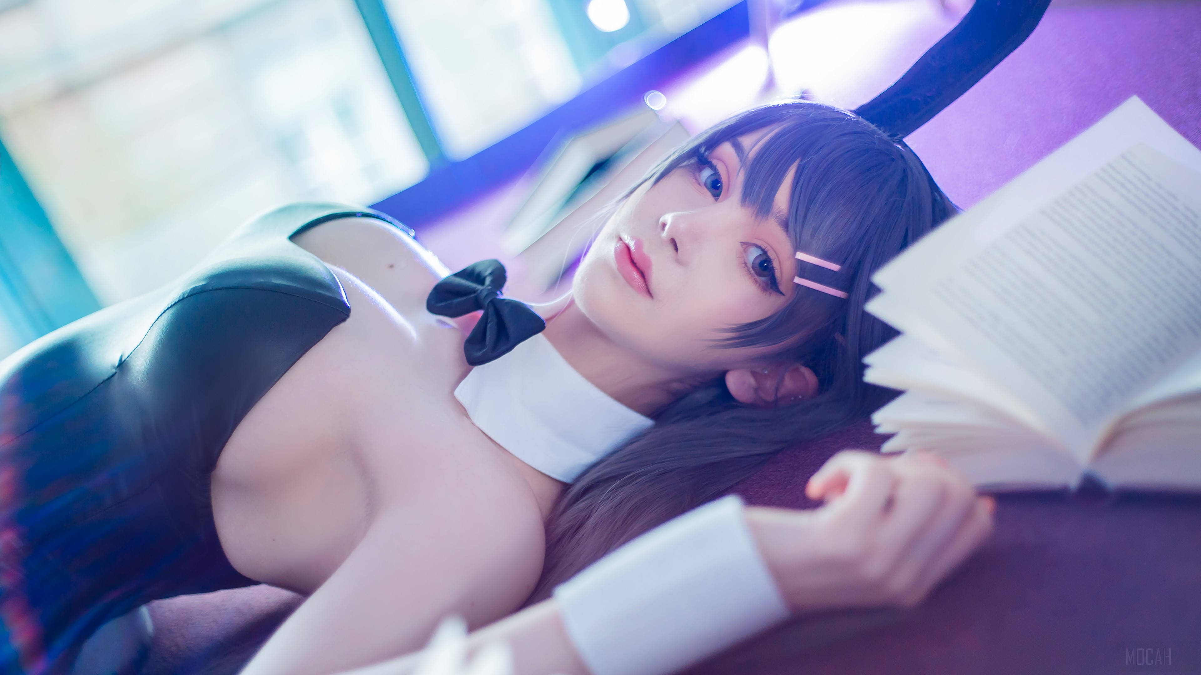 Mai 桌布 壁紙 — 一位黑髮貓耳cosplayer在柔和的紫色和藍色燈光下，懶洋洋地躺在沙發上，手裡拿著一本打開的書。
