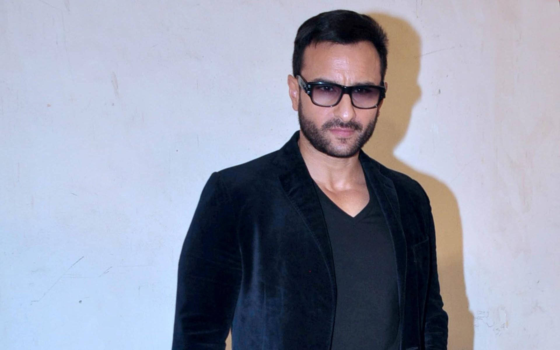 Saif Ali Khan 桌布 壁紙 — 一位黑髮鬍鬚男士，戴著時尚黑色太陽眼鏡和天鵝絨西裝外套，靠著純米白色牆壁。
