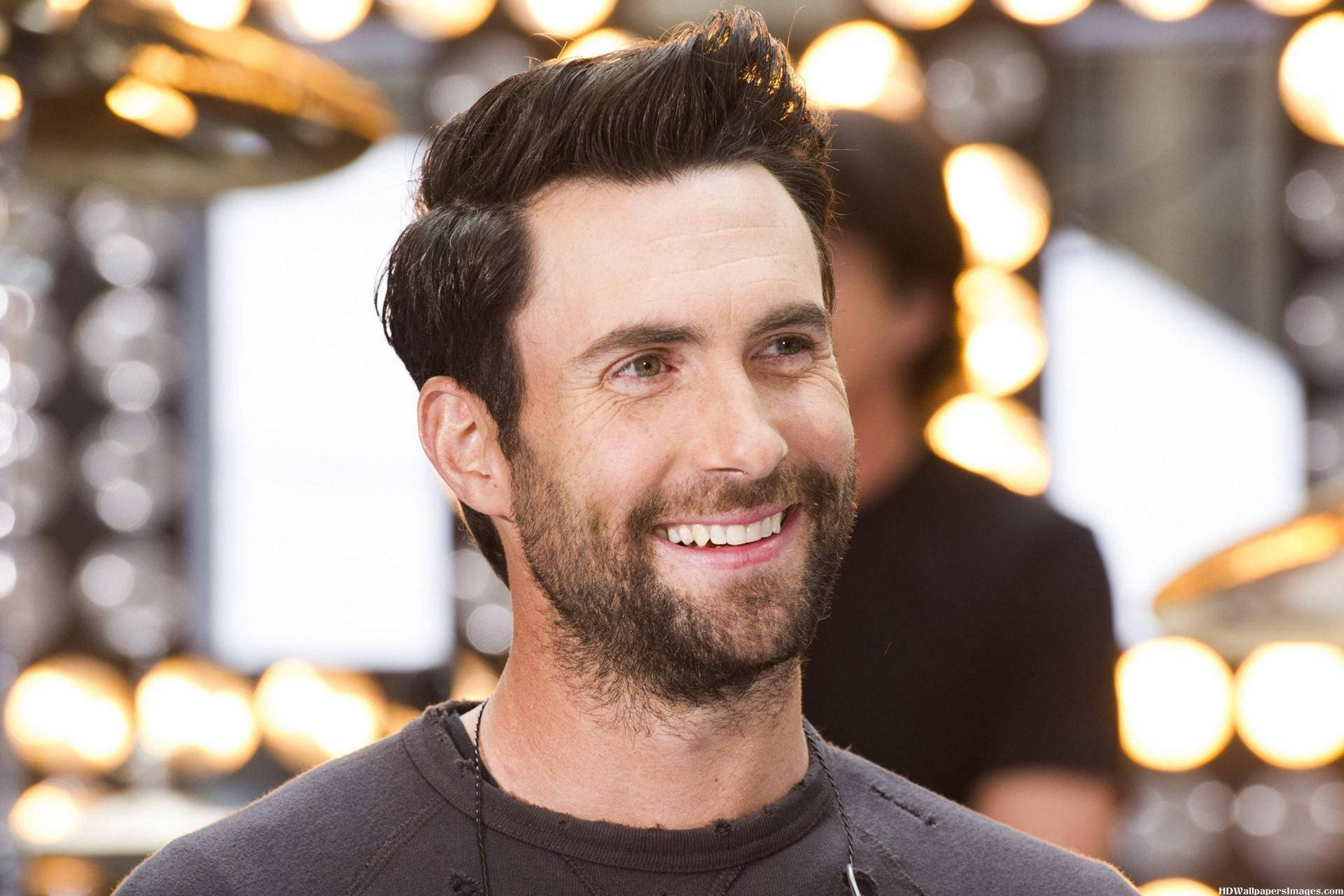 Adam Levine 桌布 壁紙 — 一位黑髮鬍鬚的男子微笑著，穿著深色襯衫，背景是發光的圓形燈光。