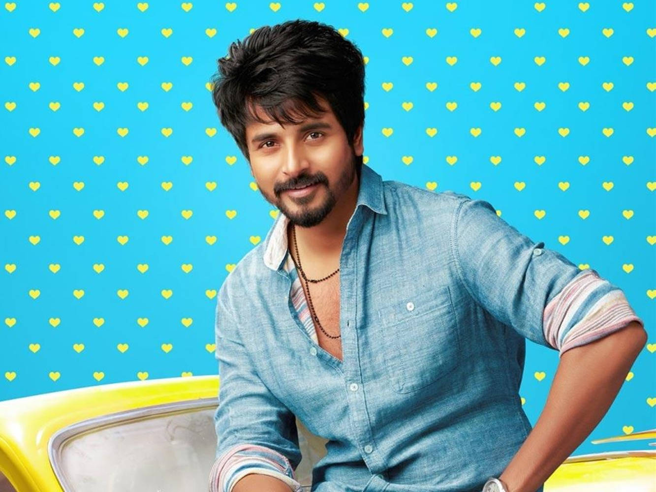 Sivakarthikeyan 桌布 壁紙 — 一位黑髮鬍鬚的男子微笑著靠在一輛黃色車上，背景是佈滿黃色心形的亮藍色牆壁。