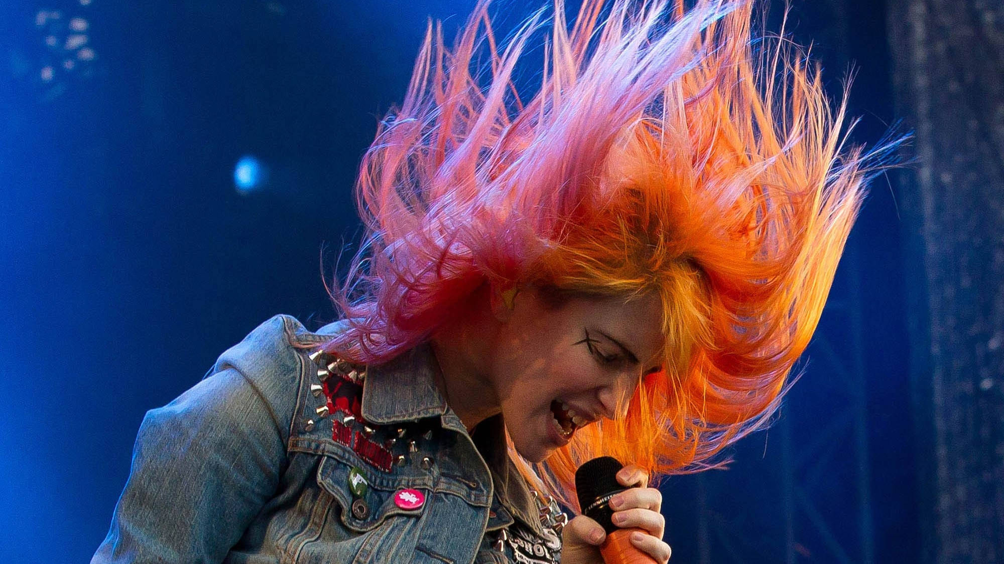 Hayley Williams 桌布 壁紙 — 一位龐克搖滾歌手，擁有充滿活力的橙色和粉紅色頭髮，在藍色舞台燈光下熱情地對著麥克風唱歌。