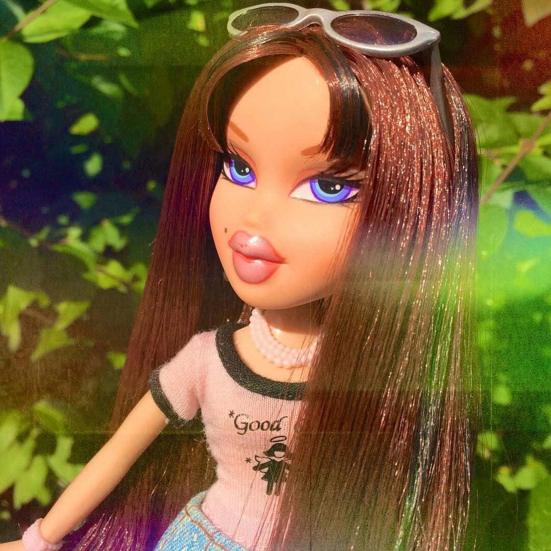bratz 美學 桌布 壁紙 — 一個 Bratz 娃娃，擁有長長的閃亮頭髮和藍色眼睛，在戶外擺姿勢，頭上戴著太陽眼鏡，背景是茂密的綠色樹葉。