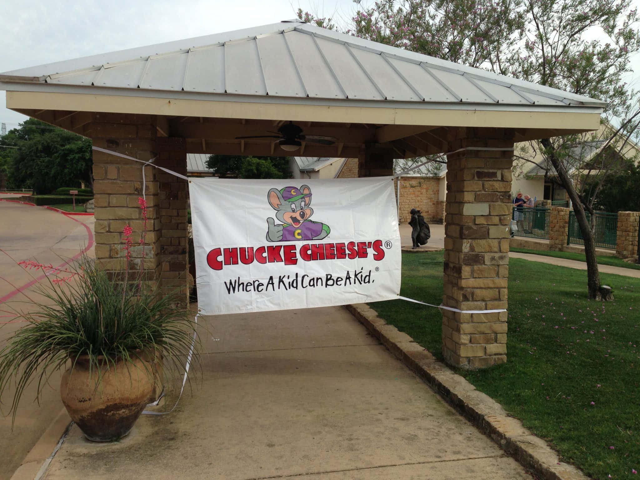 查克芝士 桌布 壁紙 — 一個 Chuck E Cheese 橫幅懸掛在金屬屋頂下的石柱之間，位於室外入口處。