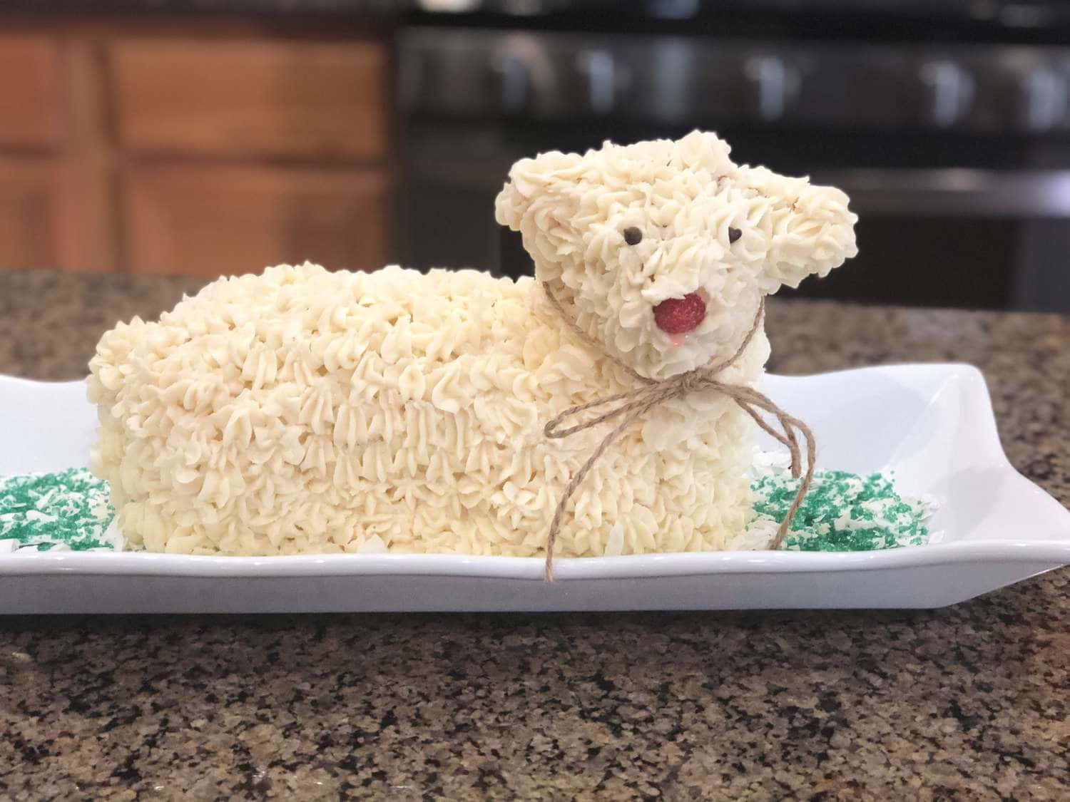 Lamb 桌布 壁紙 — 一個 fluffy 白色糖霜蛋糕，造型像羊，有紅色糖果鼻子和麻繩蝴蝶結，放在一個有綠色糖粒的白色盤子上。