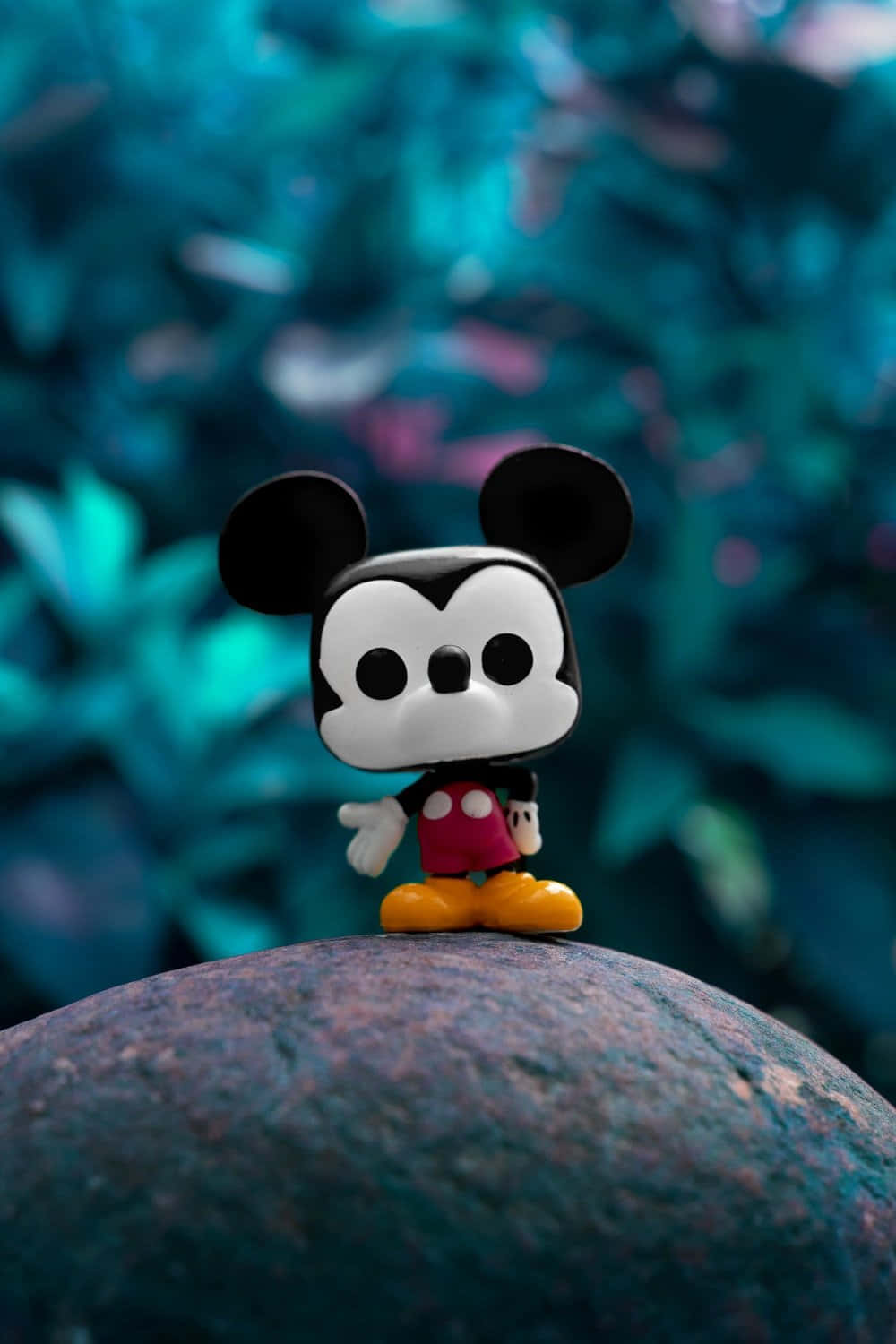 米奇 桌布 壁紙 — 一個 Funko Pop 米奇老鼠公仔站在一塊長滿苔蘚的石頭上，背景是充滿活力的藍綠色模糊樹葉。