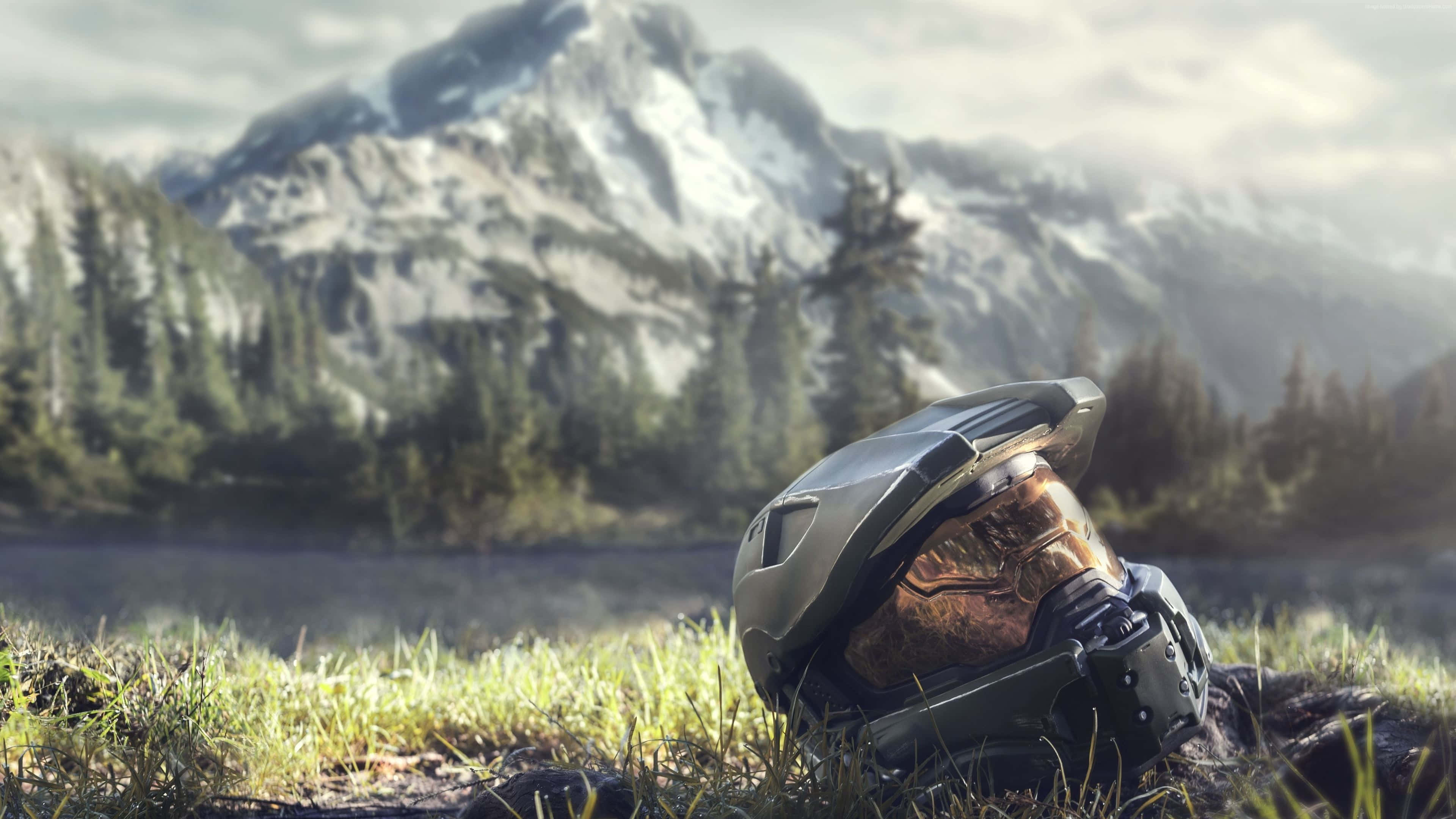 光環 桌布 壁紙 — 一個 Halo Master Chief 頭盔放在草地上，前面是霧氣瀰漫的山間湖泊，有積雪的山峰和常綠樹木。