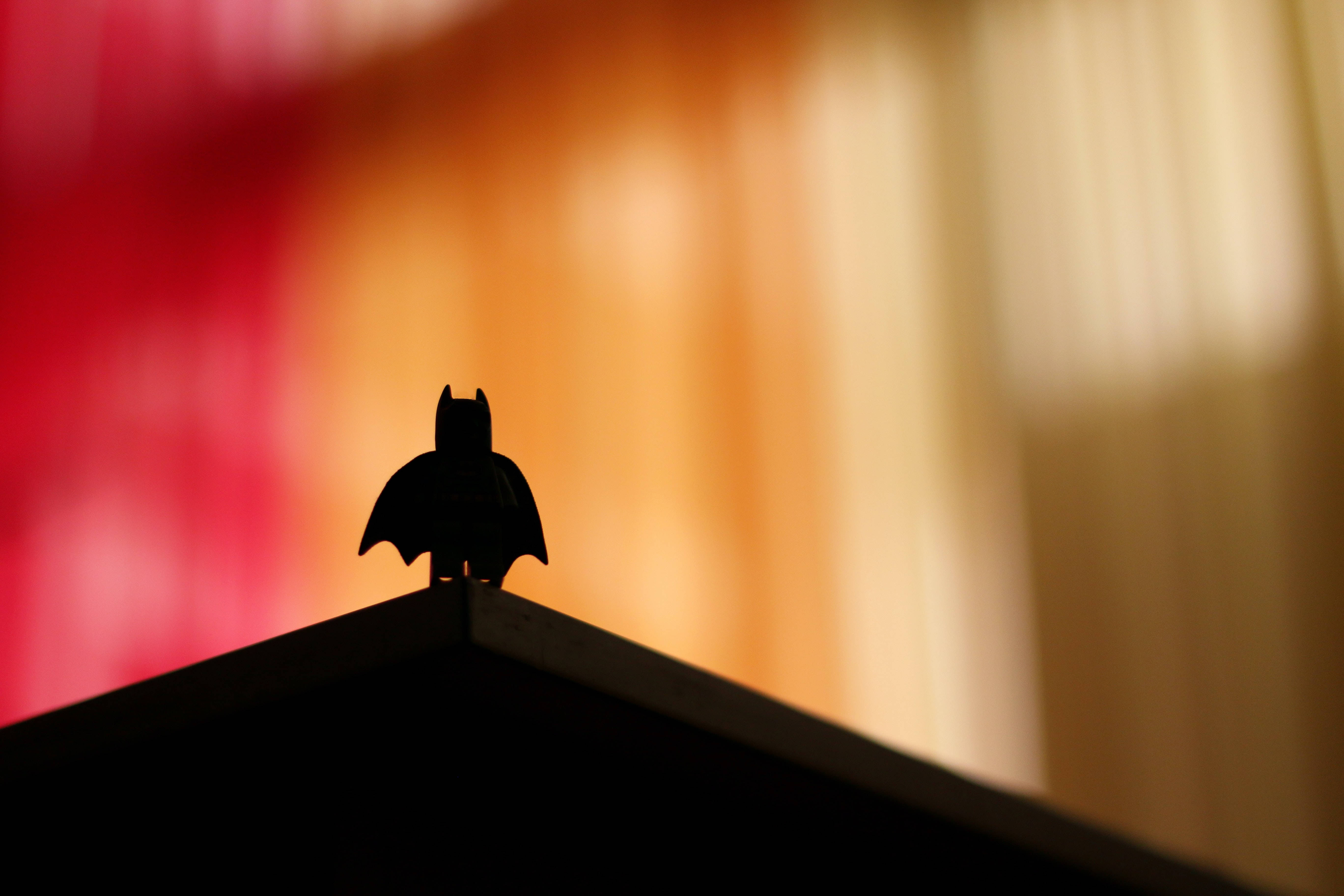 superhero supreme 桌布 壁紙 — 一個 LEGO Batman 人偶剪影棲息在屋頂頂峰上，背景是溫暖模糊的紅色和橙色色調。