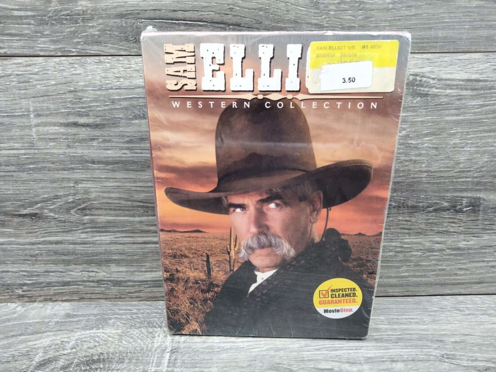 電影動圖 桌布 壁紙 — 一個 Sam Elliott Western Collection DVD 盒直立放在經風化的灰色木質表面上，特寫鏡頭顯示 Elliott 戴著牛仔帽，背景是沙漠日落。